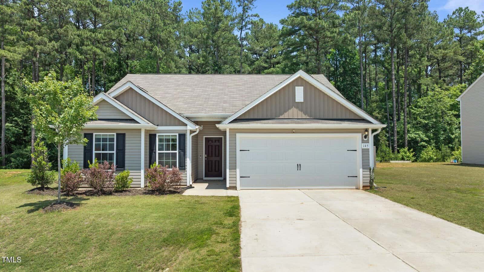 Single-story suburban house with gray siding, white trim, and a two-car garage. There are small shrubs in the front yard, a concrete driveway, and a backdrop of tall pine trees. The sky is clear and blue.