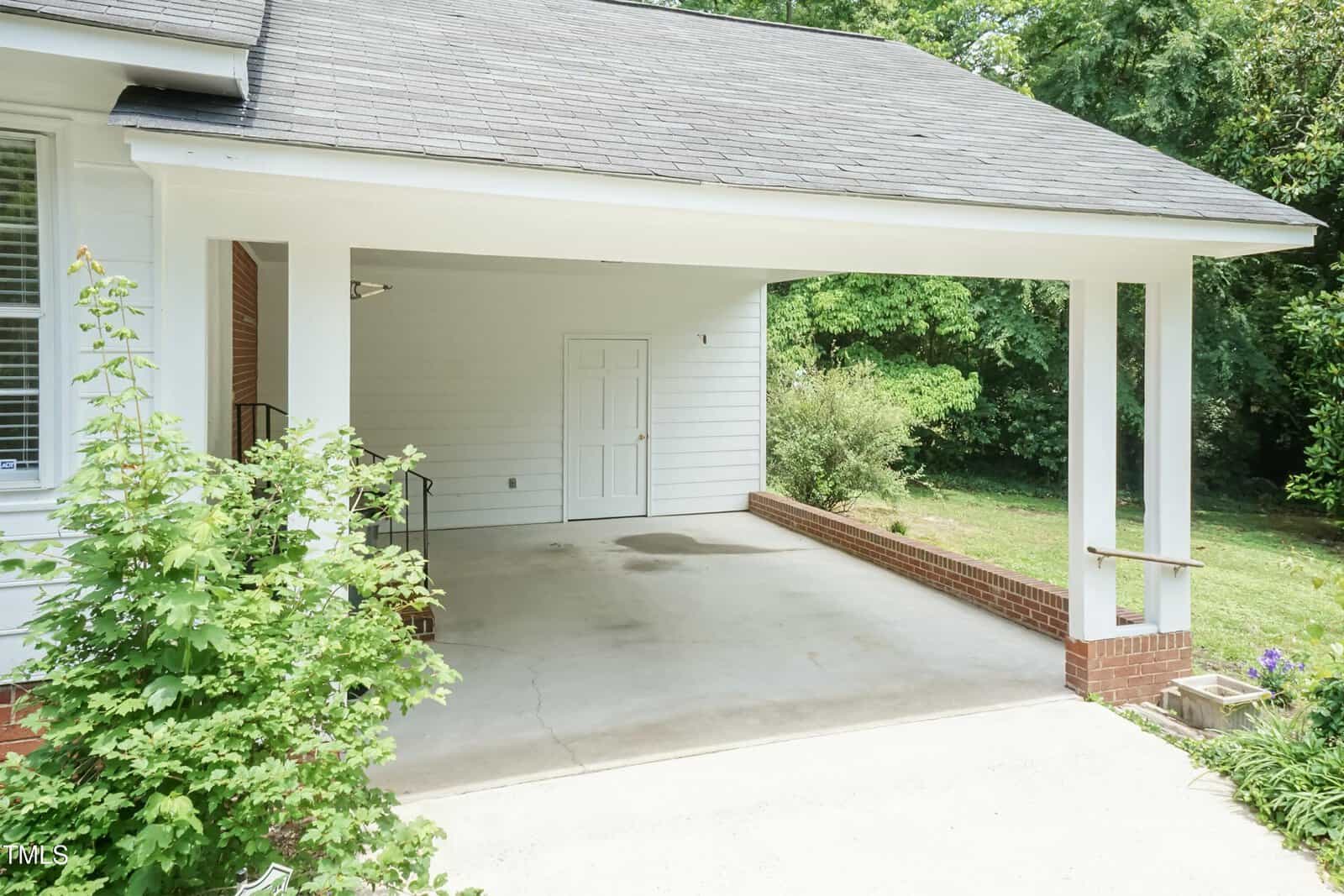 A covered carport attached to a house with white siding and brick trim. There is a white door at the back, a small metal handrail, and greenery partially visible on the left and right sides.