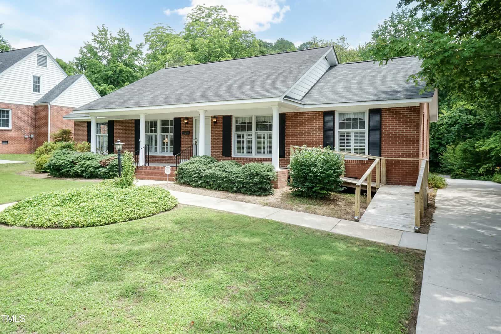 Single-story red brick house with a covered front porch, black shutters, and a wheelchair ramp leading to the entrance. The home is surrounded by green lawn, shrubs, and trees, with a paved driveway on the right.