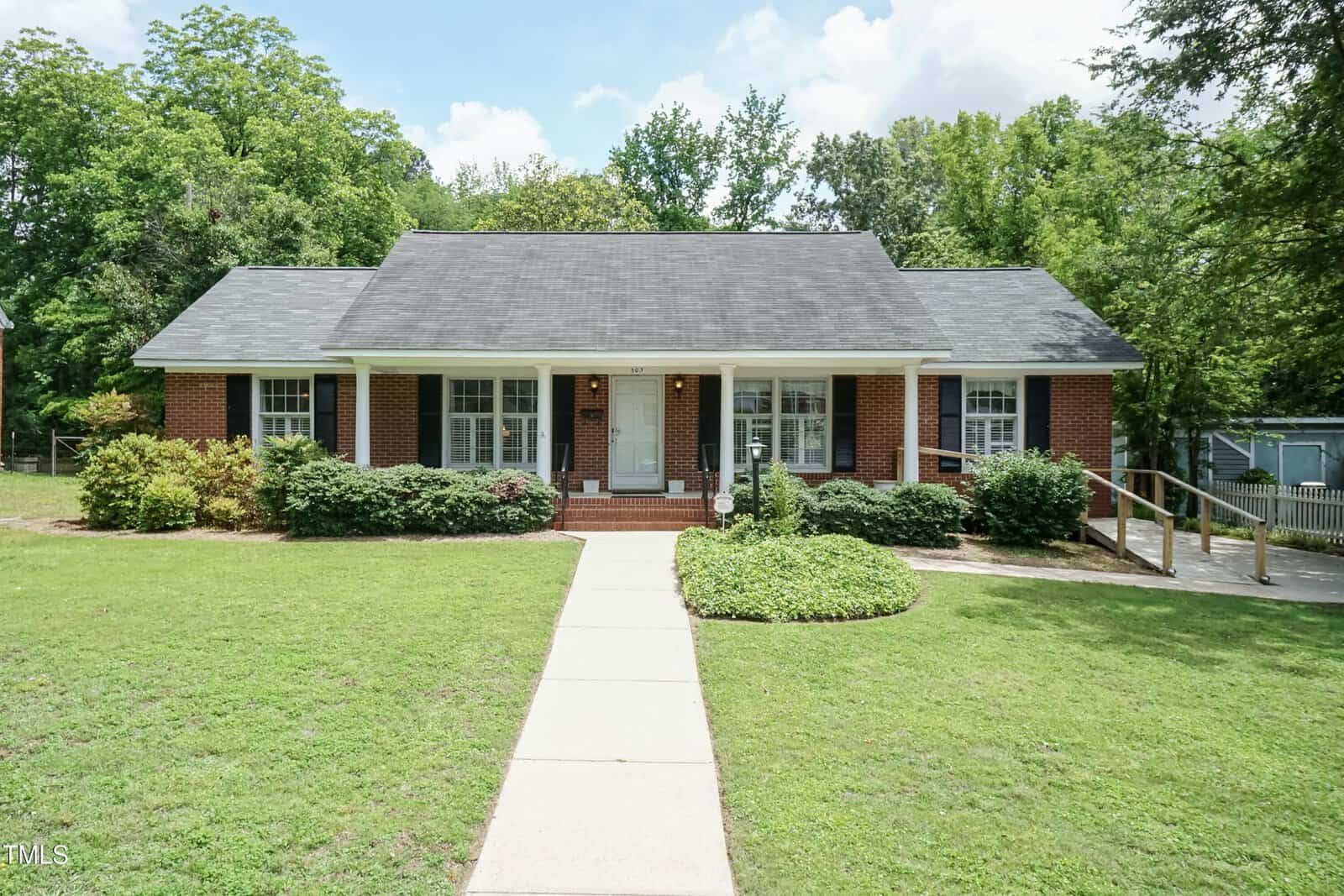 A single-story red brick house with white trim, large windows, a dark shingled roof, and a central front door, surrounded by shrubs and green lawn, with a ramp on the right side and trees in the background.