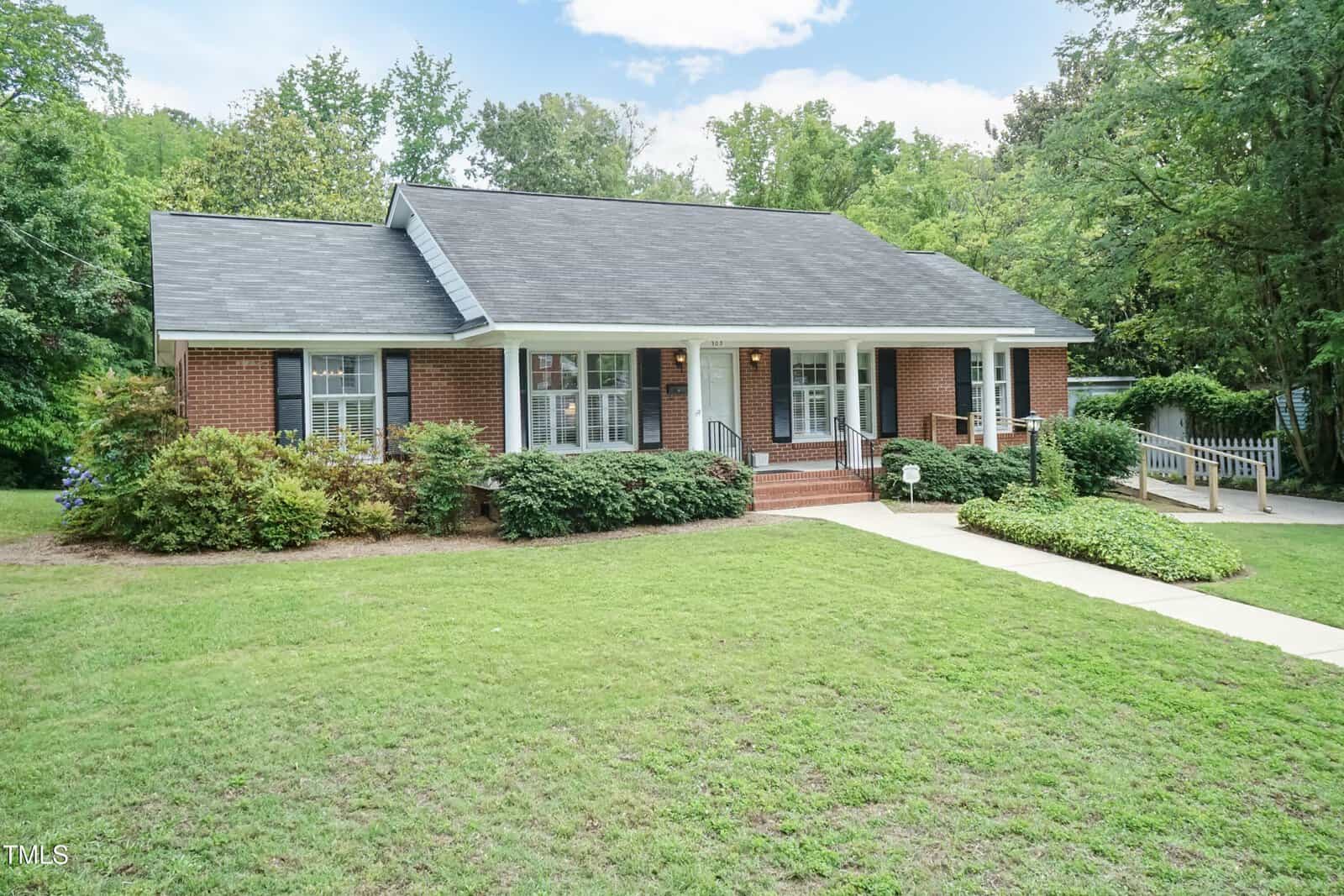 A one-story brick house with a covered front porch, surrounded by shrubs and trees, featuring a sloped lawn, front steps, and an accessibility ramp leading to the entrance.