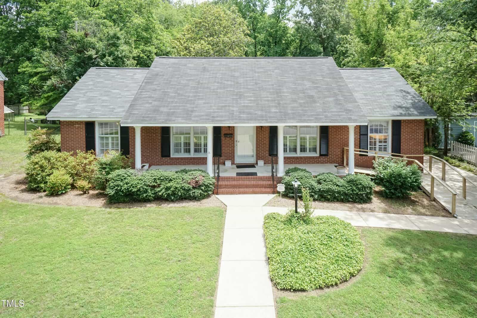 A single-story red brick house with a gray shingle roof, white trim, front porch, bushes along the front, a curved sidewalk, and a wooden accessibility ramp on the right side, surrounded by green lawn and trees.