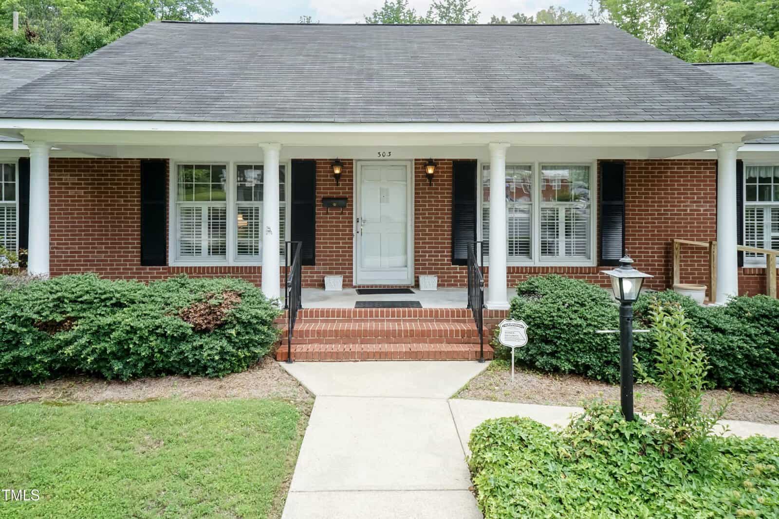 A brick house with white columns, black shutters, and a covered front porch. Bushes line the walkway leading to the front door. A lamp post and security sign are in the yard.
