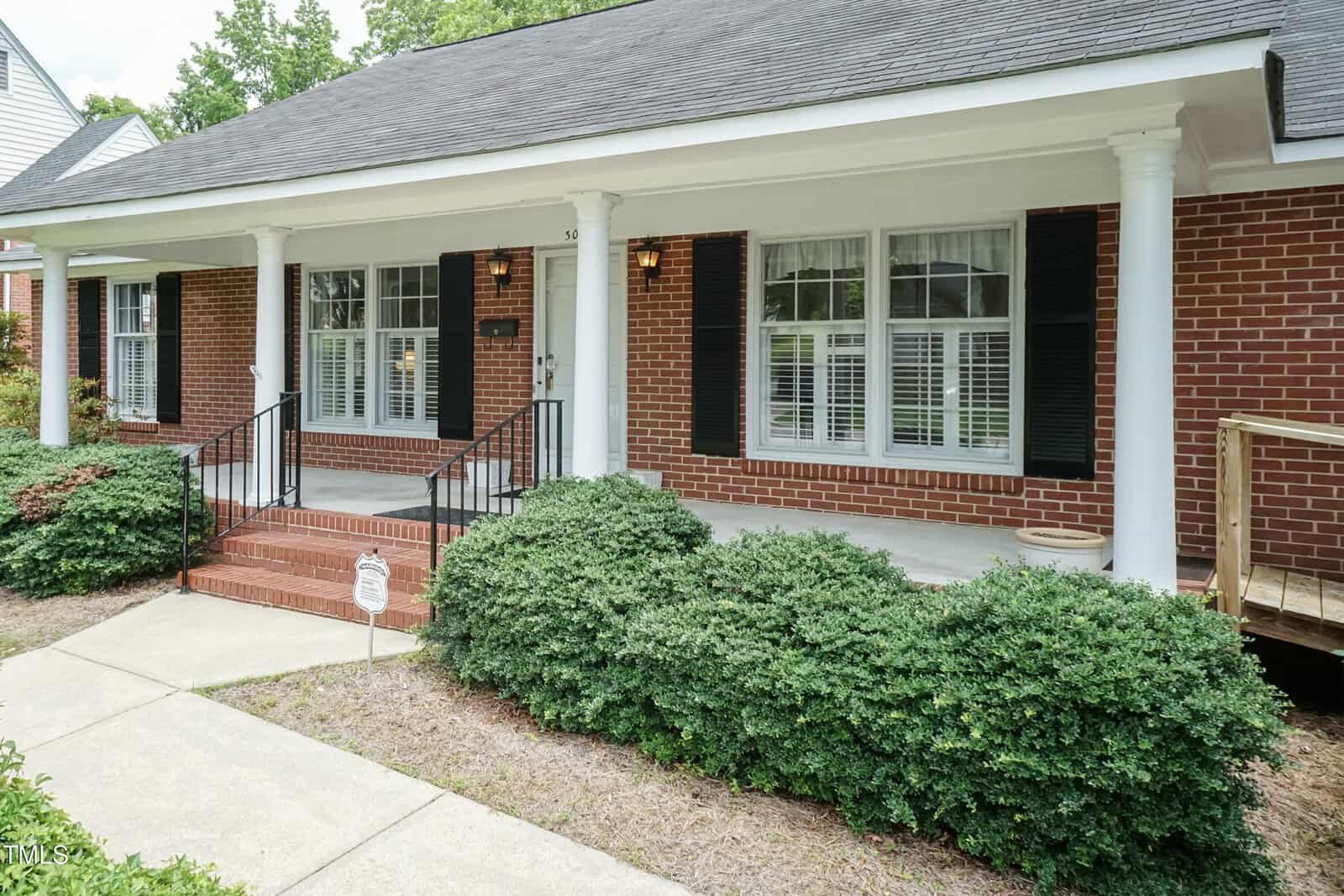 A brick house with a covered front porch, white columns, black shutters, large windows, and neatly trimmed bushes along the walkway leading to the front steps.