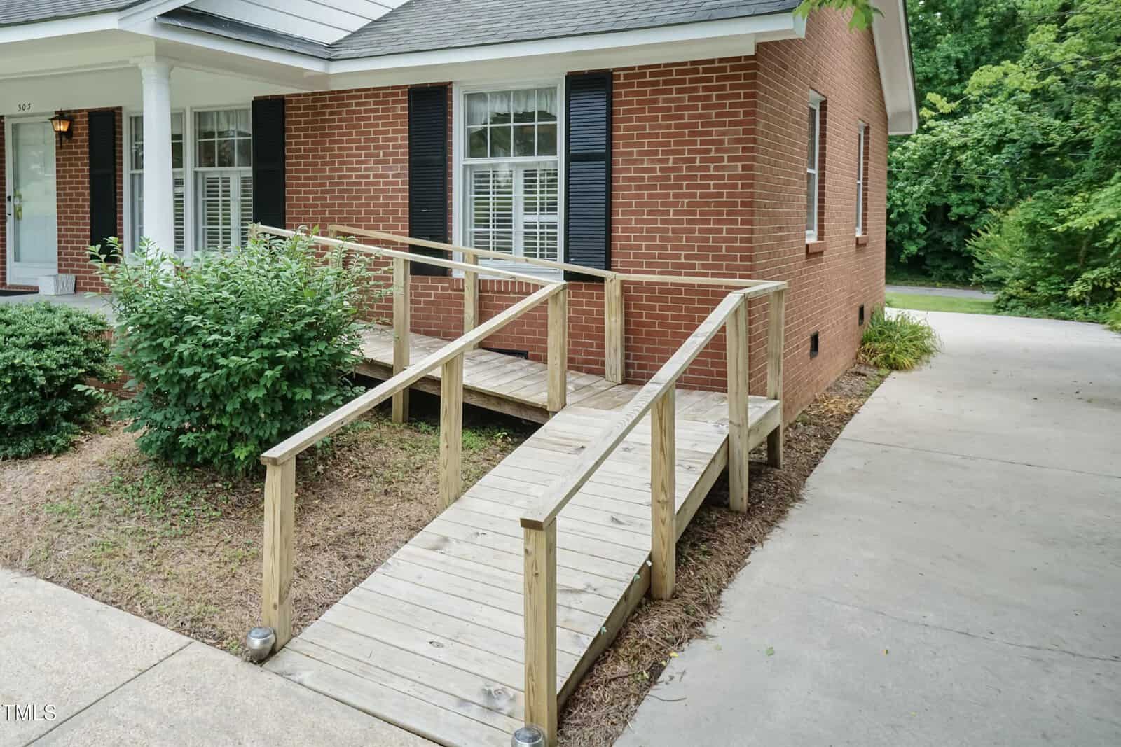 A wooden wheelchair ramp with railings leads to the front entrance of a red brick house with black shutters, adjacent to a concrete driveway and green shrubs.