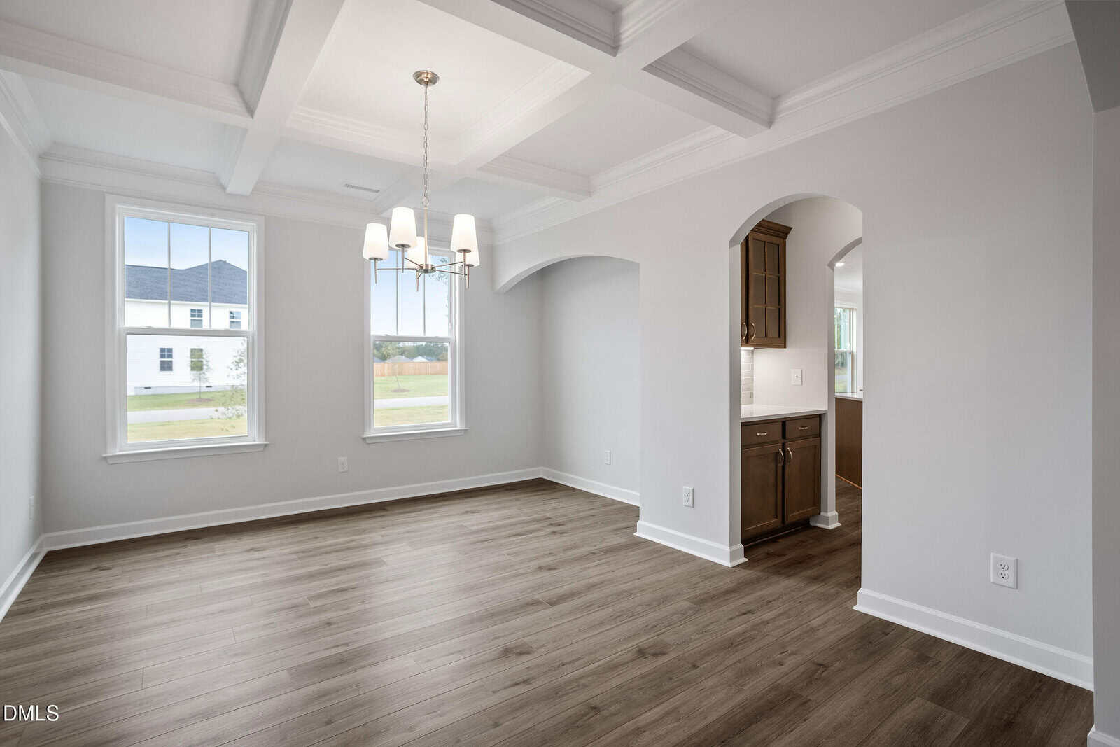 Bright, empty dining room with coffered ceiling, chandelier, large windows, light gray walls, and wood flooring. An arched doorway leads to a kitchen with brown cabinets.