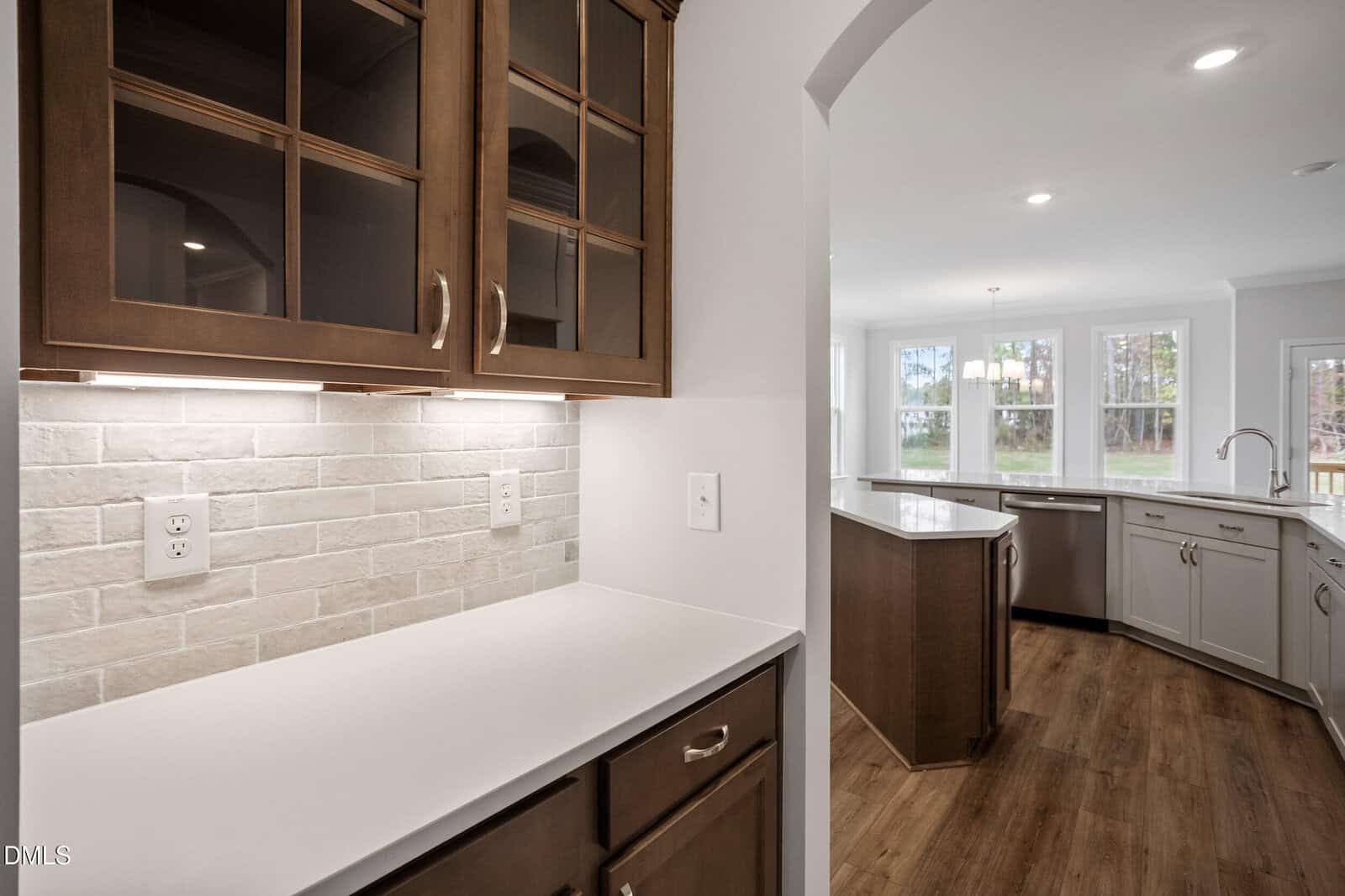 A modern kitchen features wood cabinets with glass doors, a white countertop, a light brick backsplash, an island, hardwood floors, and large windows that let in natural light.
