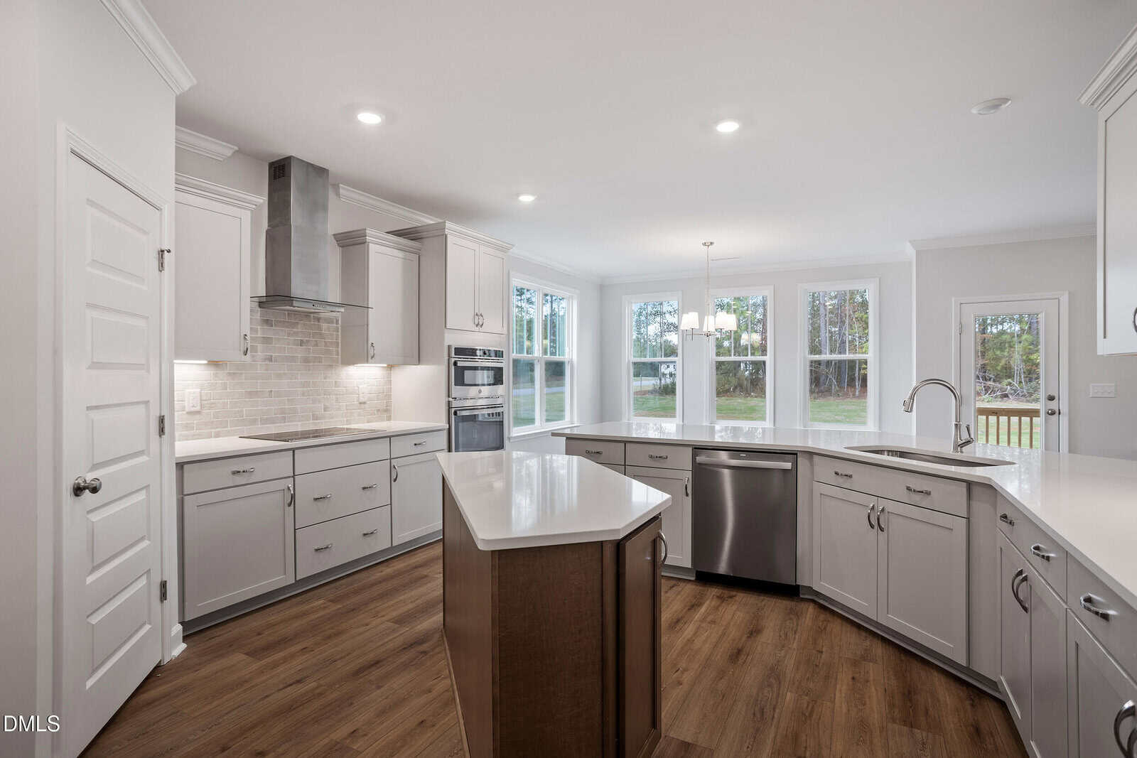 Modern kitchen with white cabinets, stainless steel appliances, a central island with a wooden base, light subway tile backsplash, wood flooring, and large windows offering a view of greenery outside.