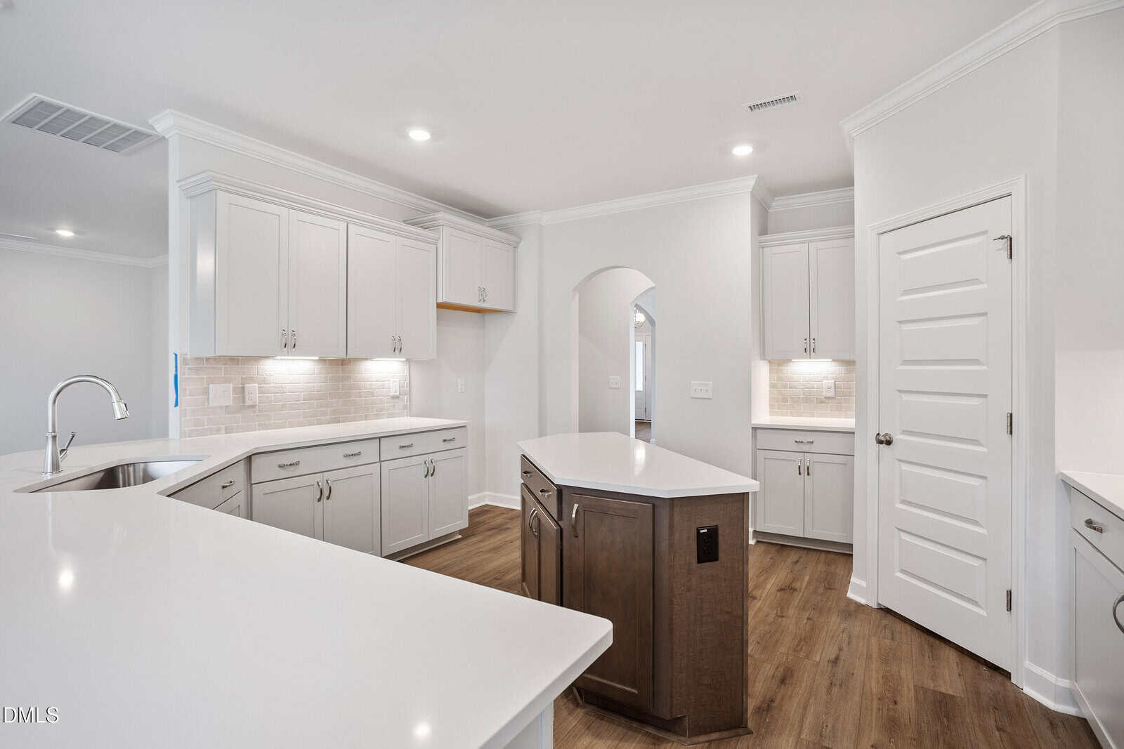 Modern kitchen with white cabinets, white countertops, a central island with dark wood base, stainless steel faucet, wood flooring, recessed lighting, and an arched doorway leading to another room.