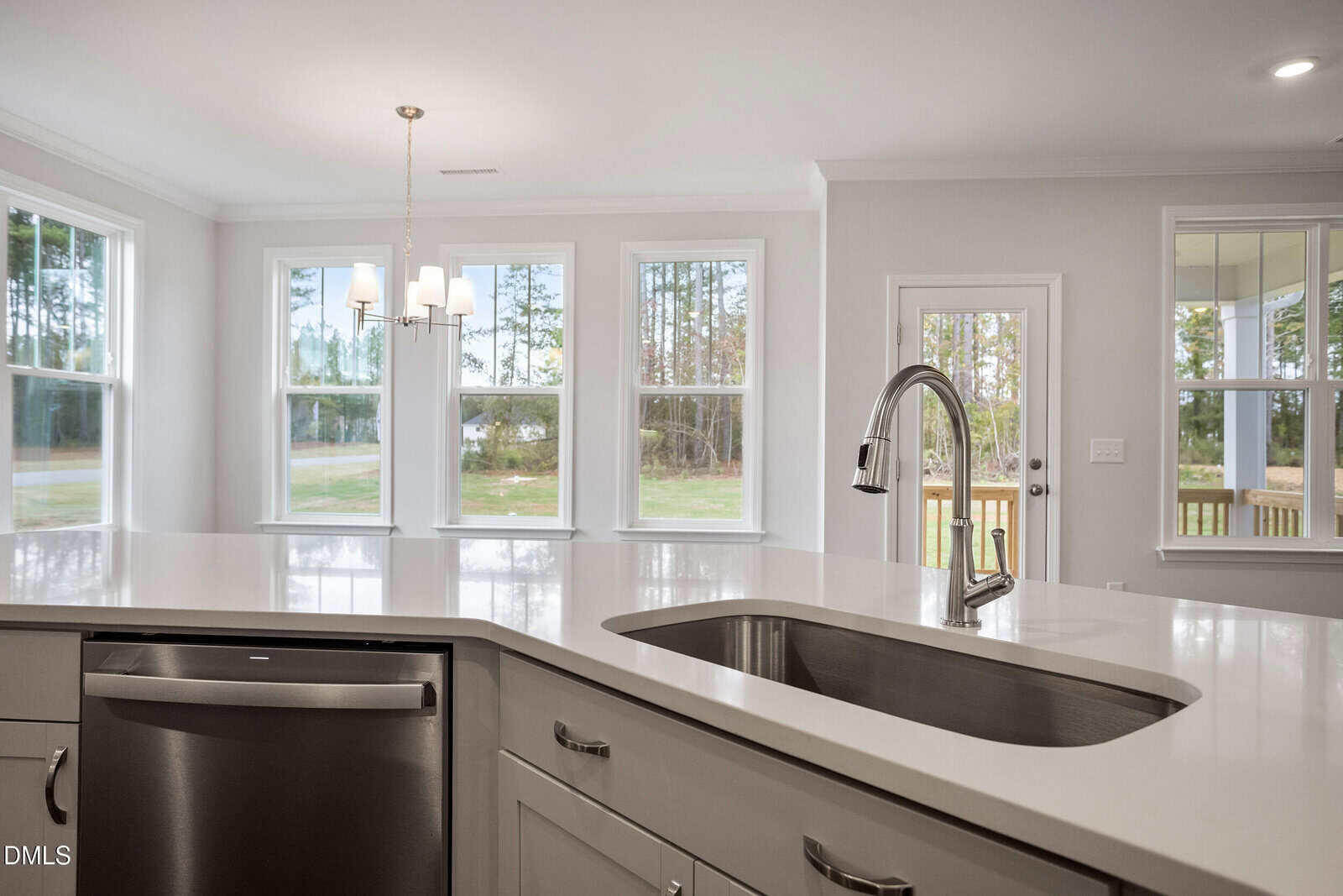 Modern kitchen with a stainless steel sink and dishwasher, white countertops, and cabinets. Large windows and a glass door in the background reveal a view of trees and a grassy yard. A light fixture hangs from the ceiling.