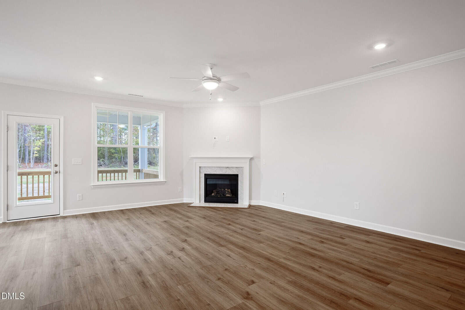 A spacious, empty living room with light-colored walls, wood flooring, a corner fireplace, large window, glass door to the outside, ceiling fan, and recessed lighting.