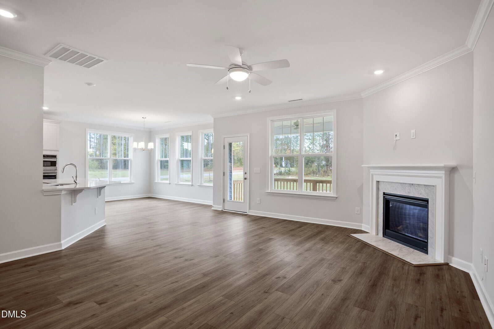 Spacious living area with light wood floors, corner fireplace, ceiling fan, large windows, and an open layout leading to a kitchen and dining space. A door and multiple windows offer views of the backyard. Walls and trim are painted white.
