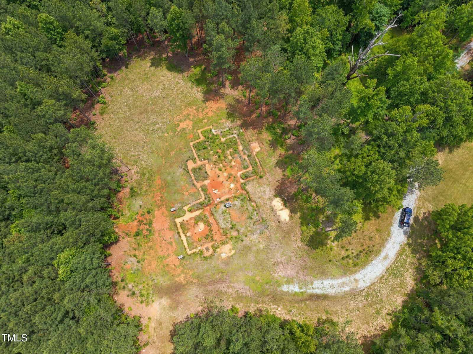 Aerial view of a forest clearing at 1627 Tommie Daniel Road, Oxford, with the foundation remains of a rectangular building. A gravel road curves nearby, leading to a parked black vehicle on the right side among green trees.