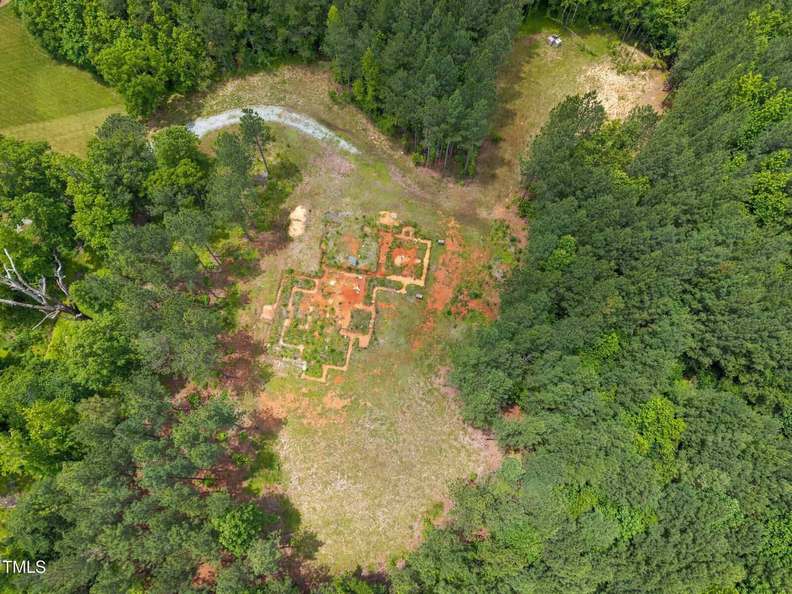 Aerial view of a forest clearing with the remnants of a building foundation, surrounded by dense trees. The structure outline is visible in the center of the image, with patches of grass and dirt.
