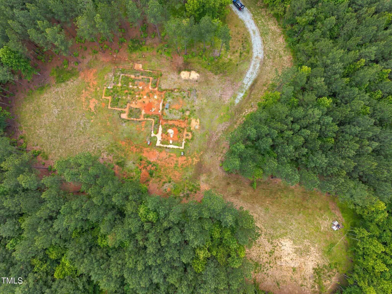 Aerial view of a forest clearing with rectangular foundations outlined in the dirt, a curved gravel road, and dense green trees surrounding the area; a parked car is visible at the top right of the image.