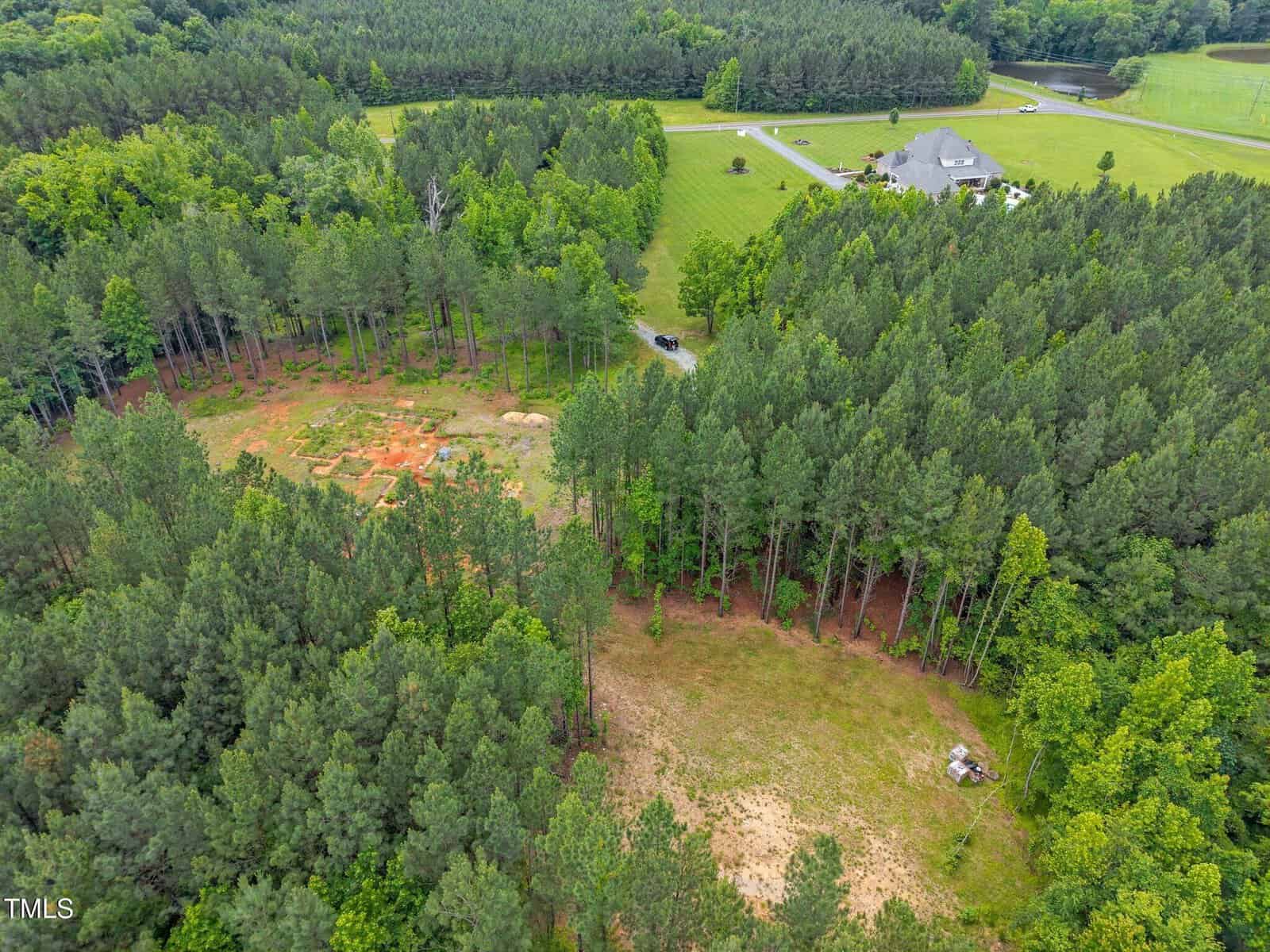 Aerial view of a wooded area with cleared patches of land, a winding driveway, and a house with a large lawn in the background, surrounded by dense trees and open fields.