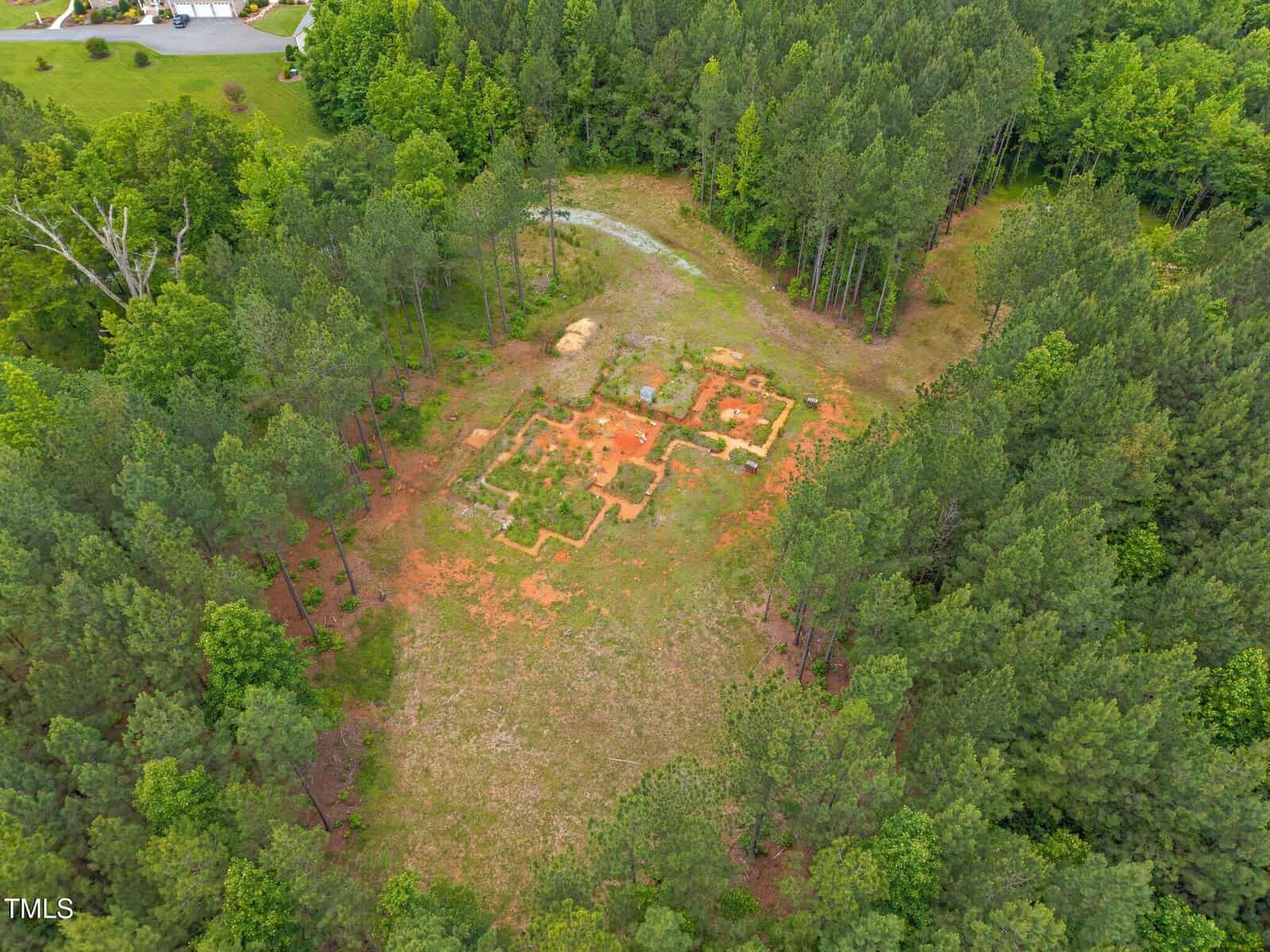 Aerial view of a forest clearing with visible rectangular foundation outlines in the soil, surrounded by dense green trees and a small dirt path leading to the site.