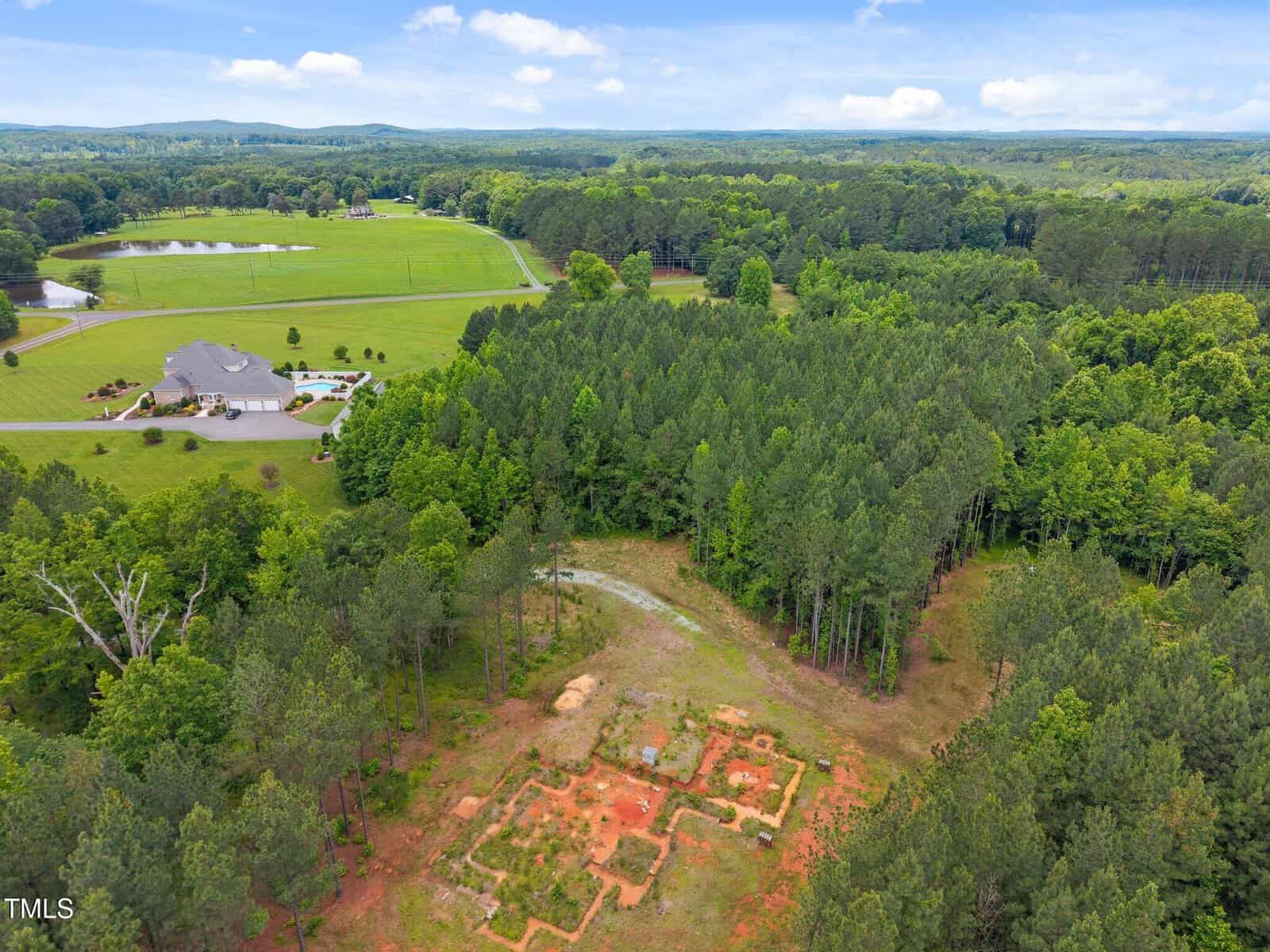 Aerial view of a wooded area with a dirt clearing containing outlined foundations, near a large house with a pool, surrounded by open grassy fields, trees, a pond, and distant forested hills under a partly cloudy sky.