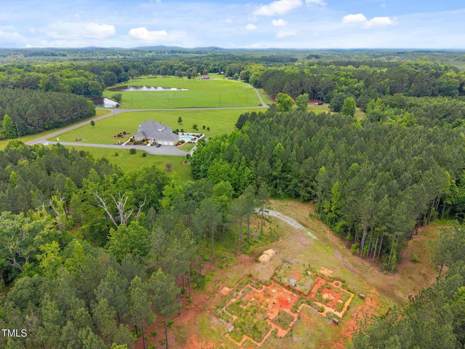 Aerial view of a large house with a pond, lawn, and driveway surrounded by fields and forest. In the foreground, red soil and partially cleared land with visible outlines of a possible foundation.