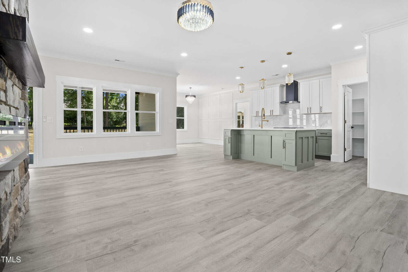Spacious modern kitchen and dining area with light wood floors, white cabinets, a large green island, pendant lights, a chandelier, and large windows letting in natural light.
