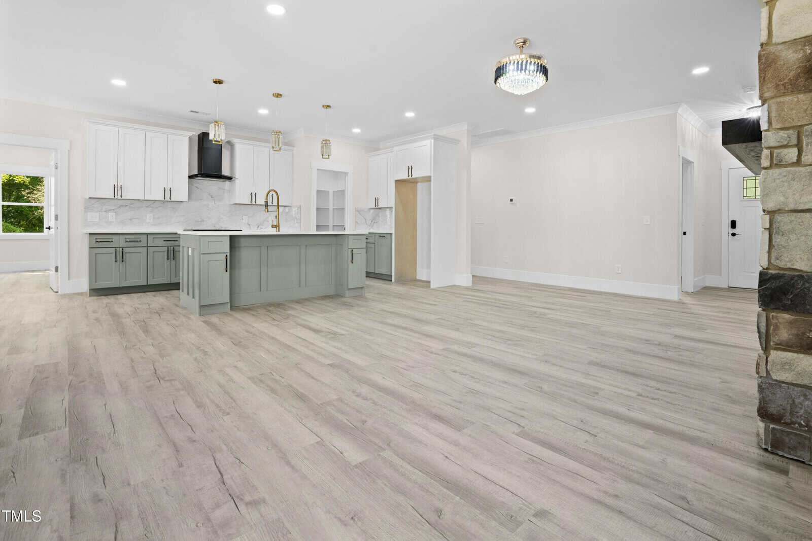 Spacious modern kitchen with light wood floors, white walls, a large gray island with pendant lights, white cabinets, marble backsplash, and gold hardware. Adjacent open area, stone fireplace on the right, and abundant natural light.