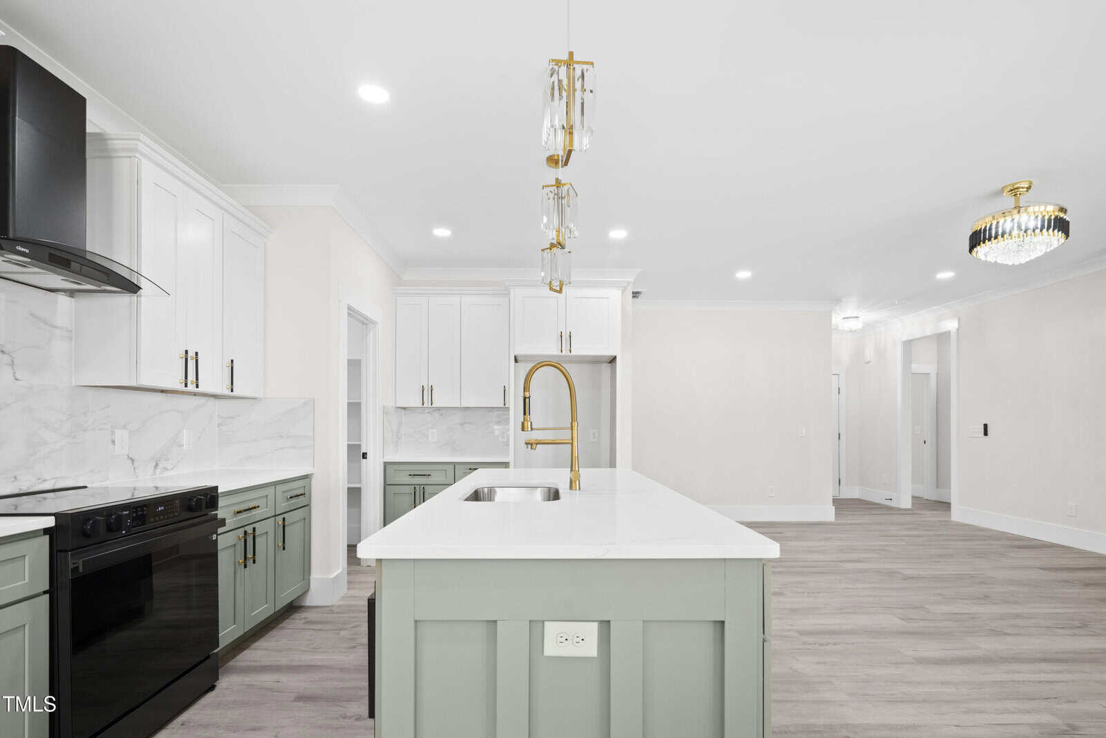 Modern kitchen with white and green cabinets, marble countertops, a center island with a gold faucet, black appliances, and two pendant lights; open layout with light wood flooring and a chandelier in the adjacent room.