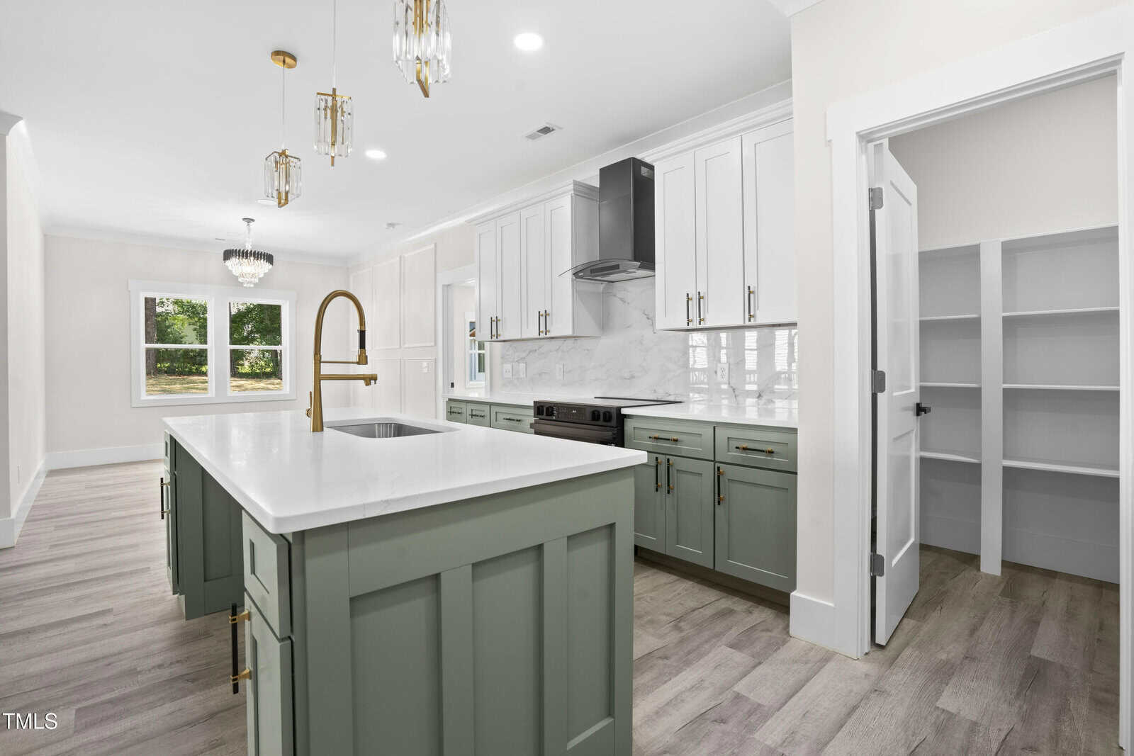 Modern kitchen with a large island, green lower cabinets, stainless steel appliances, gold faucet, pendant lights, white upper cabinets, marble backsplash, light wood flooring, and an open pantry door.