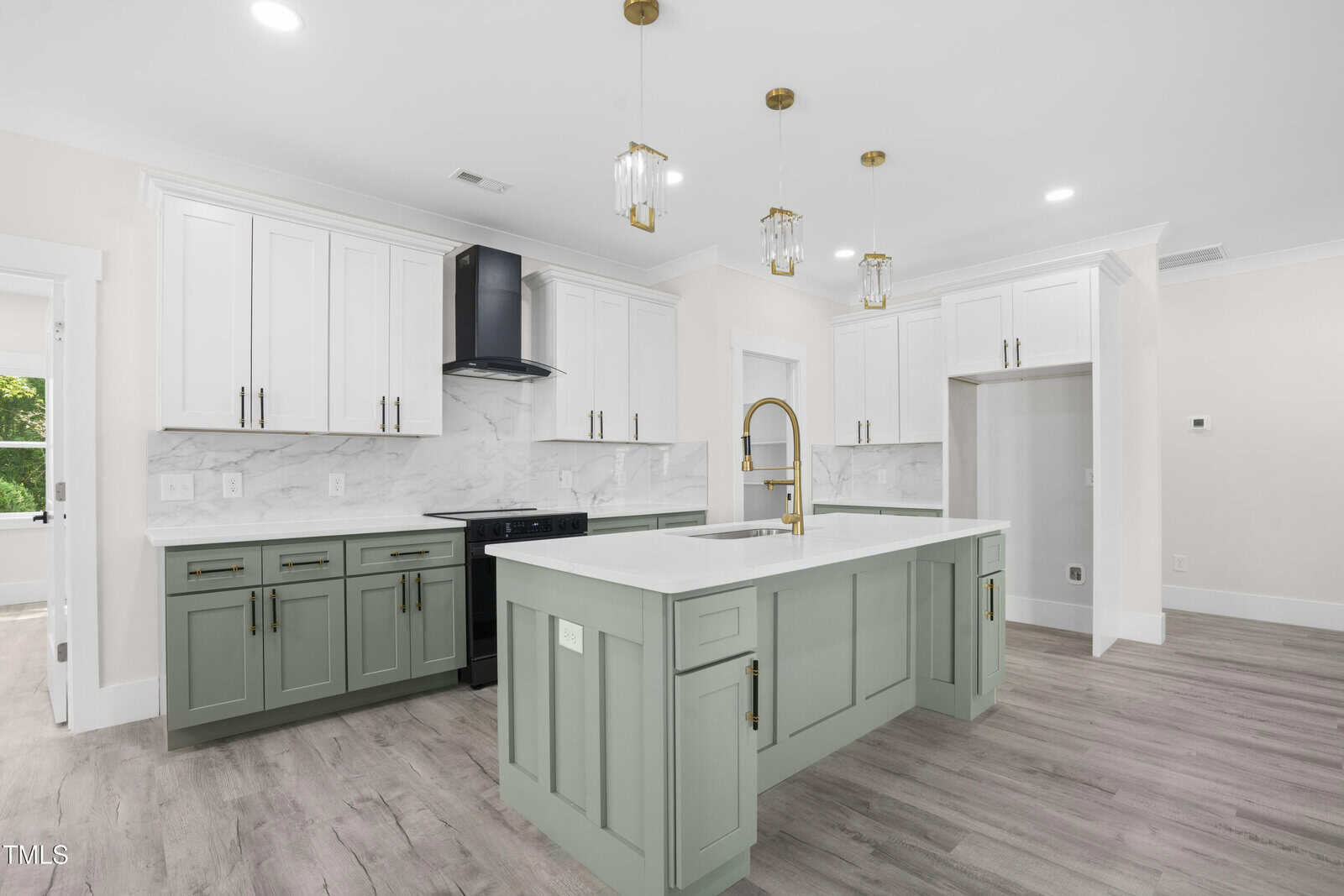 Modern kitchen with white and sage green cabinets, marble backsplash, gold hardware, pendant lights, a black range, center island with sink, and light wood flooring. White walls and trim complete the space.