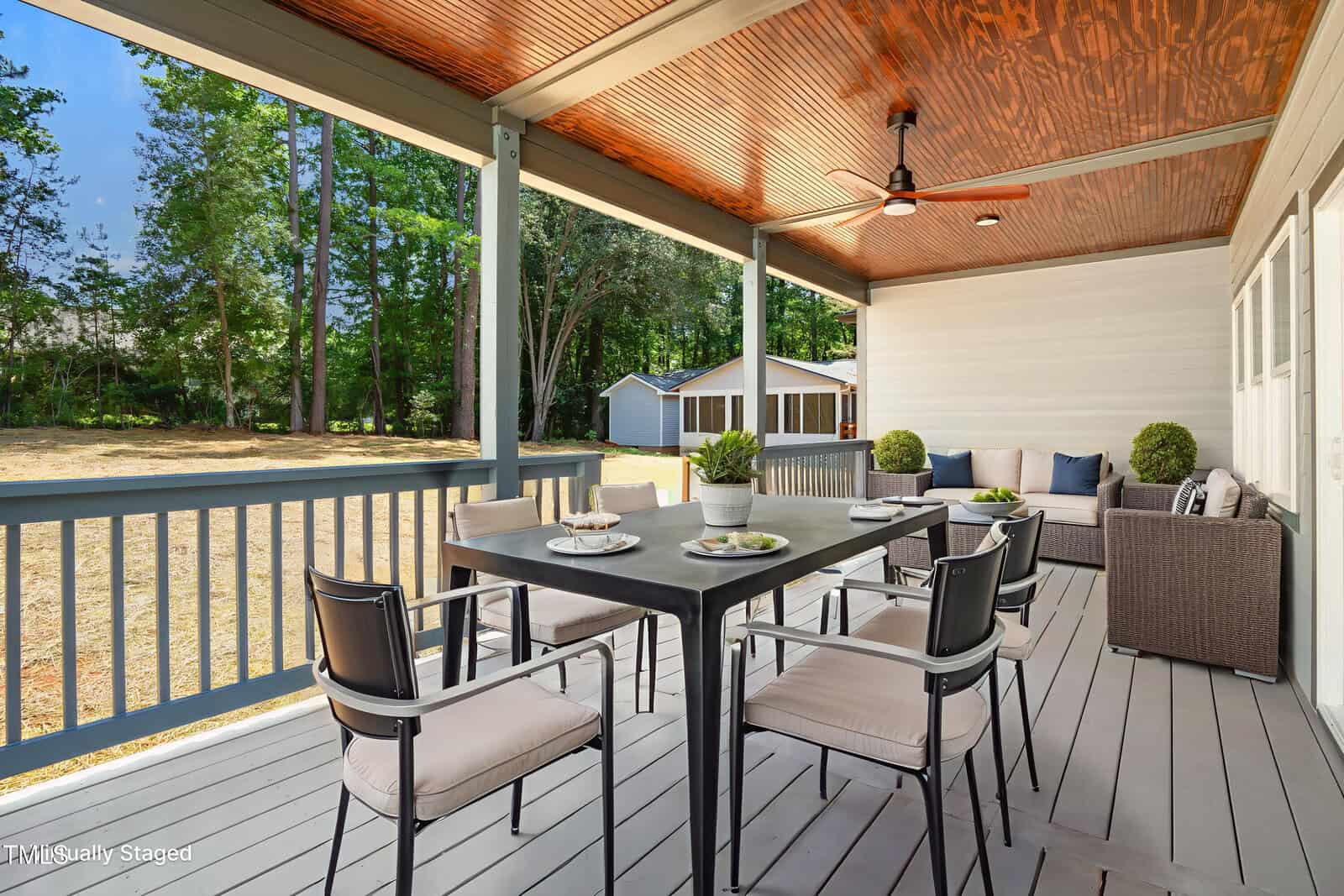A covered porch with a ceiling fan, modern patio furniture, a black dining table set for four, and a sofa seating area overlooks a spacious backyard with trees and an outbuilding in the background.