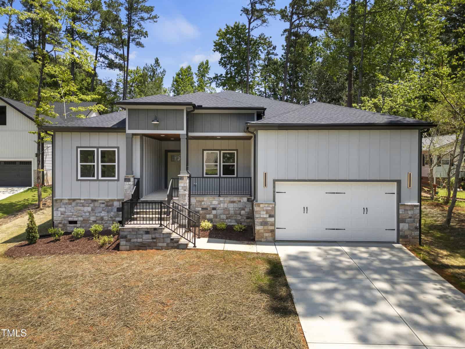 Modern single-story house with gray siding, stone accents, and black trim. A covered front porch, steps with metal railings, and a double garage are visible. Surrounded by trees and landscaped yard.
