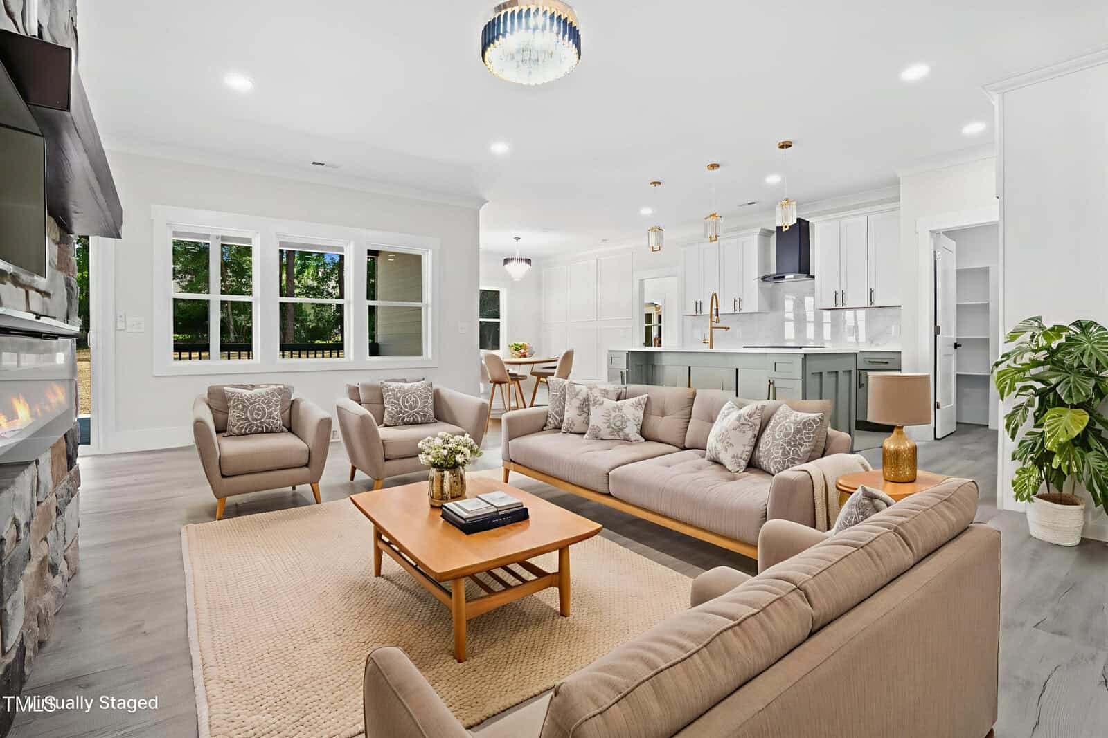 A bright, modern open-concept living room and kitchen with beige sofas, armchairs, a wooden coffee table, and a stone fireplace. The kitchen features white cabinets, an island, and gold pendant lights.
