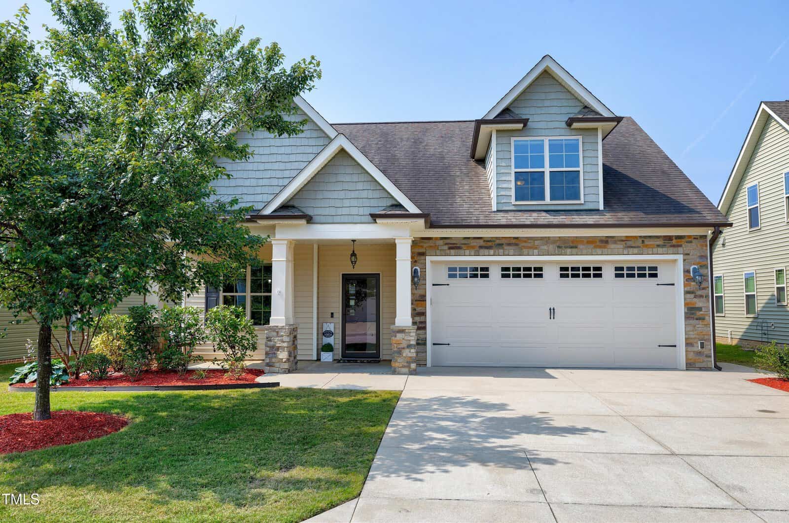 A two-story suburban house with a stone and siding exterior, a covered front porch, large windows, and a double garage. The yard is landscaped with a tree, grass, and red mulch around the plants.