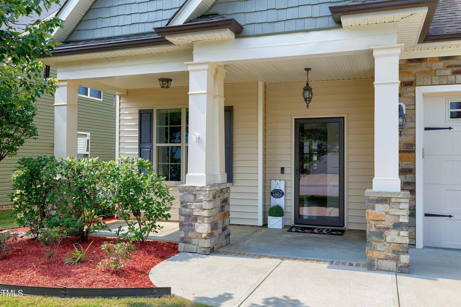A modern house entrance with beige siding, stone pillars, a glass front door, hanging lantern, covered porch, and landscaped garden beds with red mulch and green shrubs.