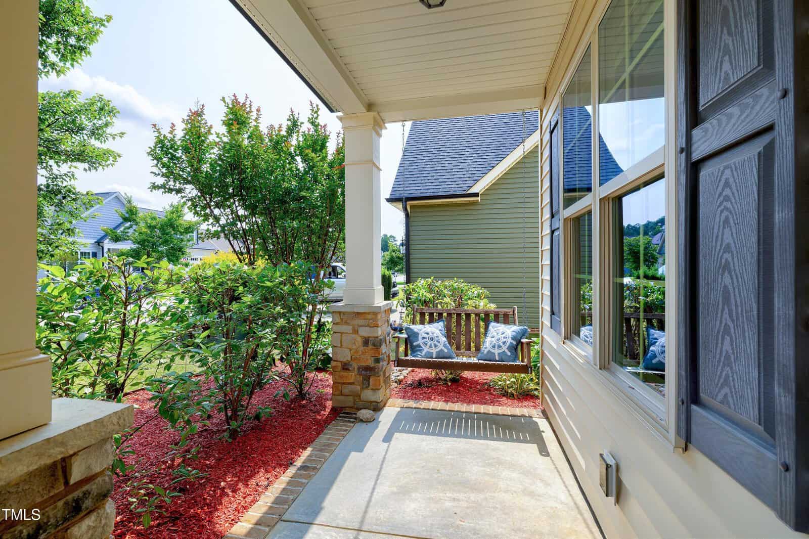 A cozy front porch with a wooden swing bench decorated with patterned pillows overlooks a well-maintained garden with green shrubs and red mulch. Sunlight brightens the scene and reflects off the front windows.
