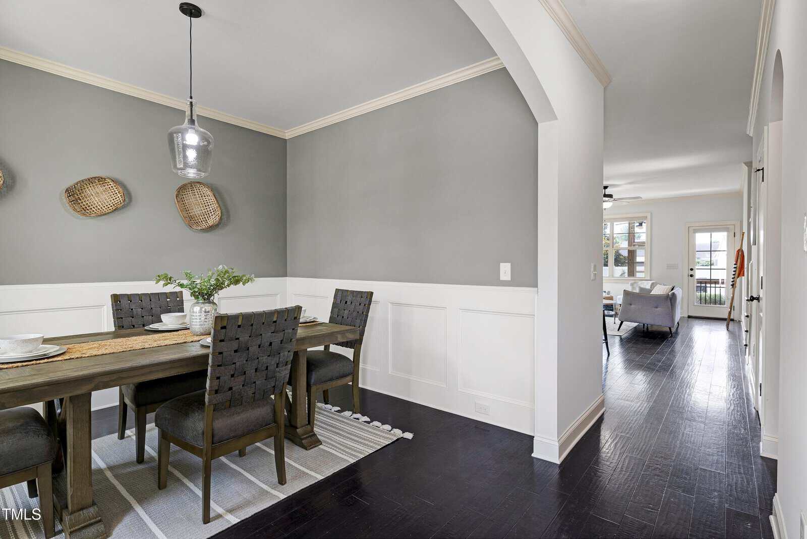 A modern dining room with a wooden table set for four, woven chairs, a pendant light, gray walls with white wainscoting, and a decorative wall with baskets. The space opens into a bright living room.