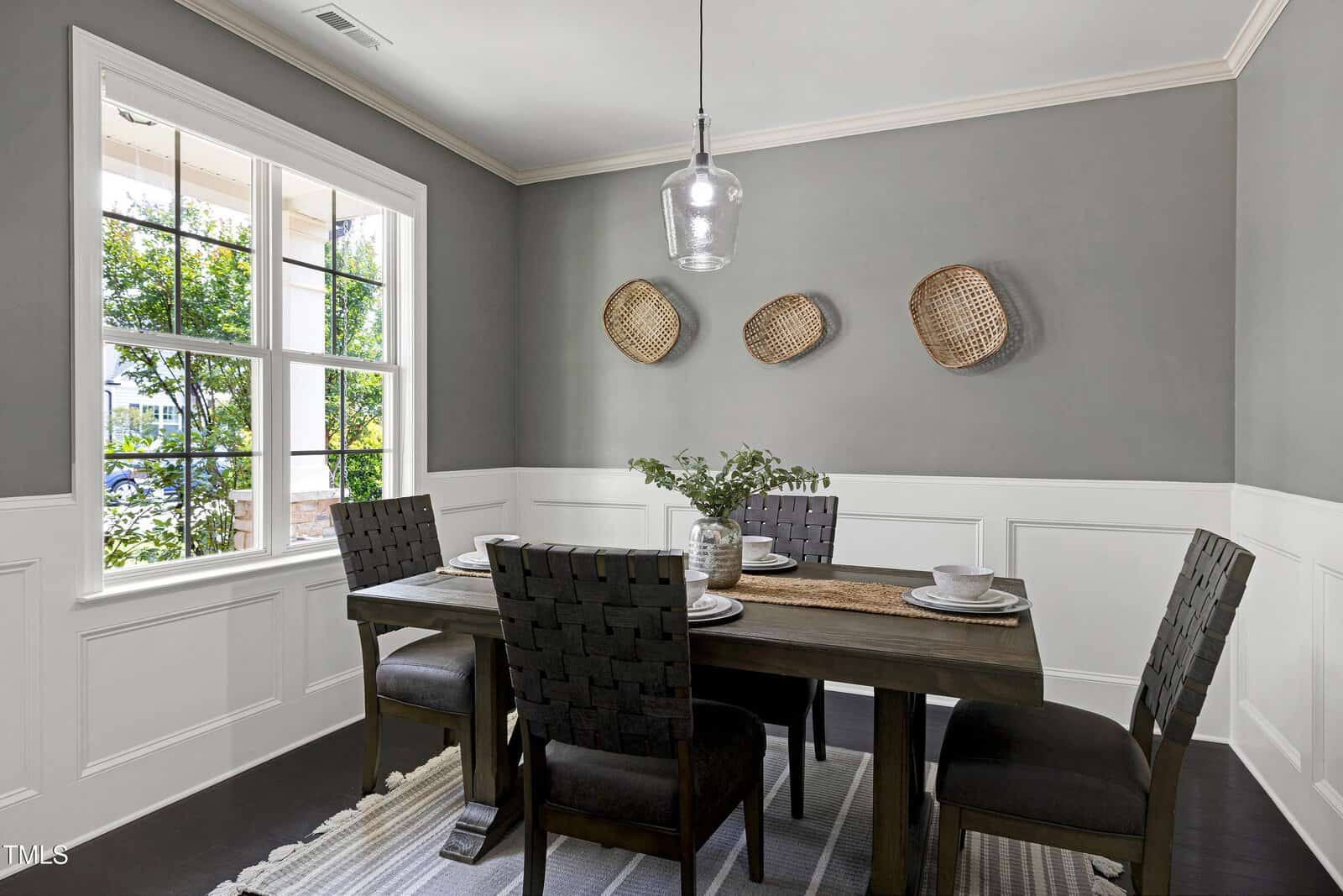 A dining room with a wooden table, four dark chairs, a woven table runner, and a glass pendant light. Three wicker baskets hang on a gray accent wall above white wainscoting. A large window lets in natural light.