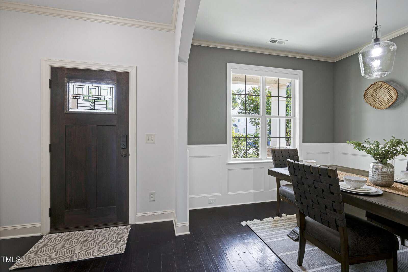 A modern entryway and dining area with dark wood floors, a black door with glass panels, a gray accent wall, a window, a dining table with chairs, and a hanging light fixture. Plants and tableware are on the table.