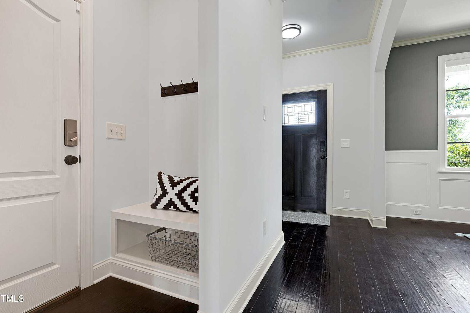 A small entryway with a white bench and black patterned pillow, a coat rack with hooks above, a wire basket below, dark wood flooring, and a front door with sidelights in the background.