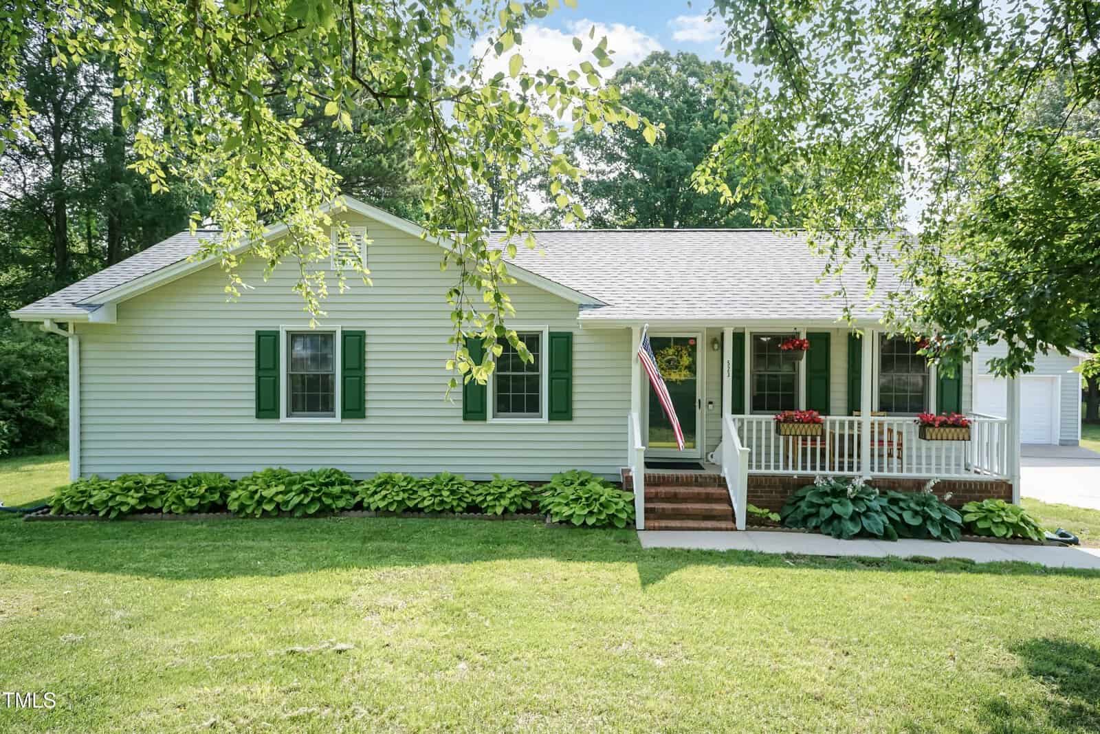 Single-story house with light green siding, green shutters, and a covered front porch. An American flag hangs by the steps, with flower boxes on the porch railing and a neatly maintained lawn in front.