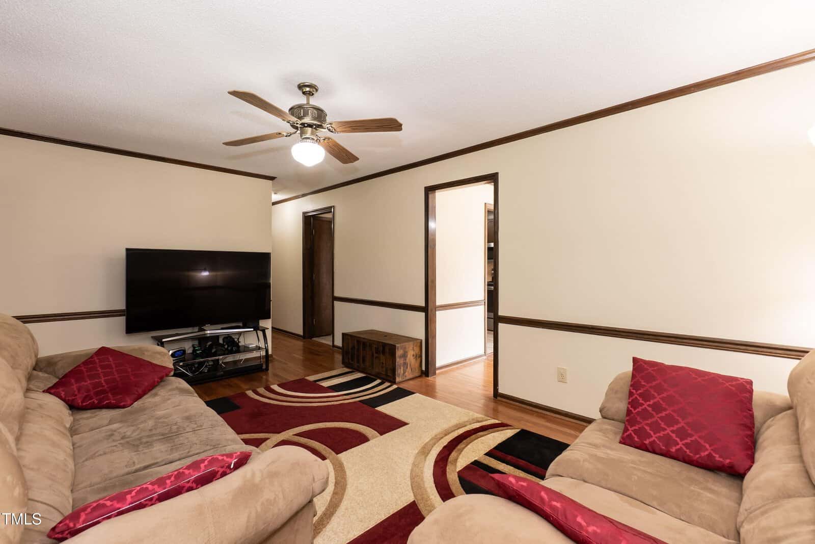 A living room with beige walls and wood trim, two tan sofas with red pillows, a patterned area rug, a flat-screen TV on a stand, a ceiling fan with lights, and hardwood flooring.