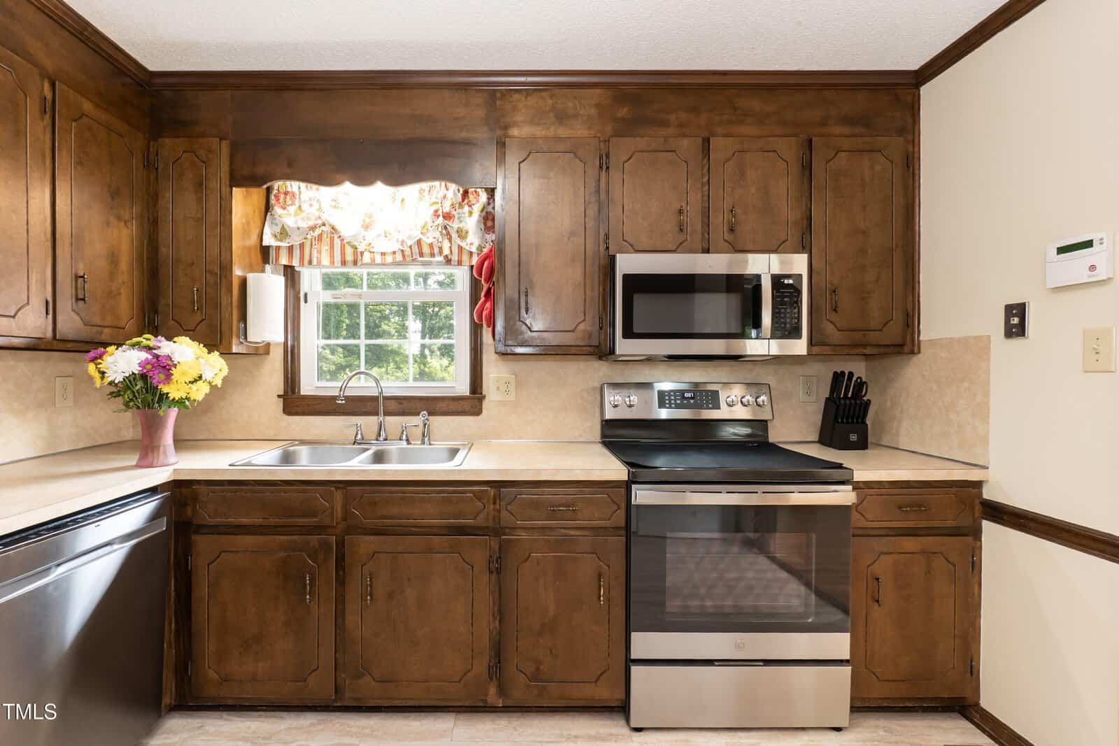 A kitchen with dark wood cabinets, a stainless steel stove and microwave, a double sink under a window with a floral valance, a bouquet of flowers on the counter, and a knife block next to the stove.