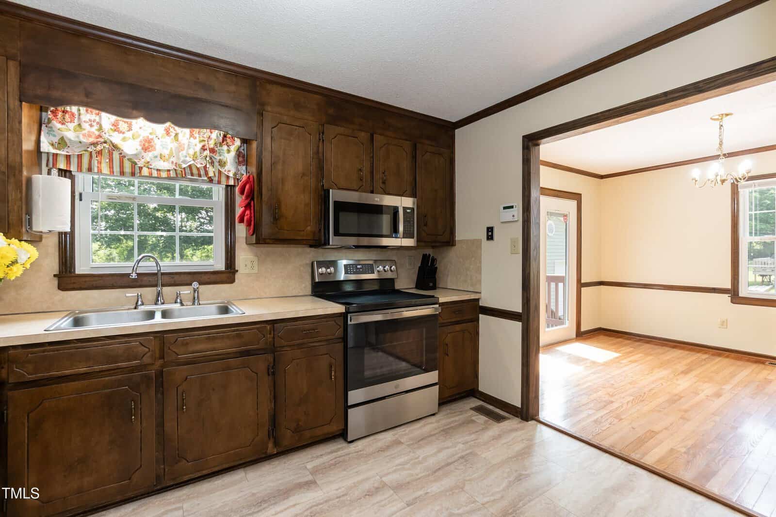 A kitchen with dark wood cabinets, beige countertops, a stainless steel stove and microwave, a double sink under a window with a floral valance, and an adjacent dining room with wood floors and a chandelier.