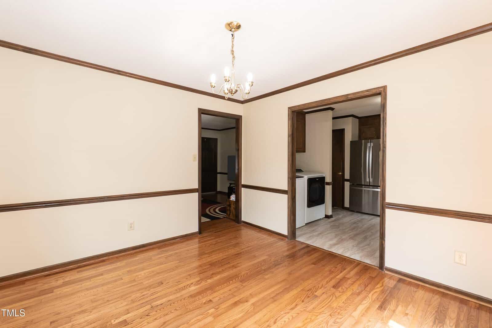 A bright room with hardwood floors, cream walls, dark wood trim, a gold chandelier, and open doorways leading to a kitchen with stainless steel appliances.