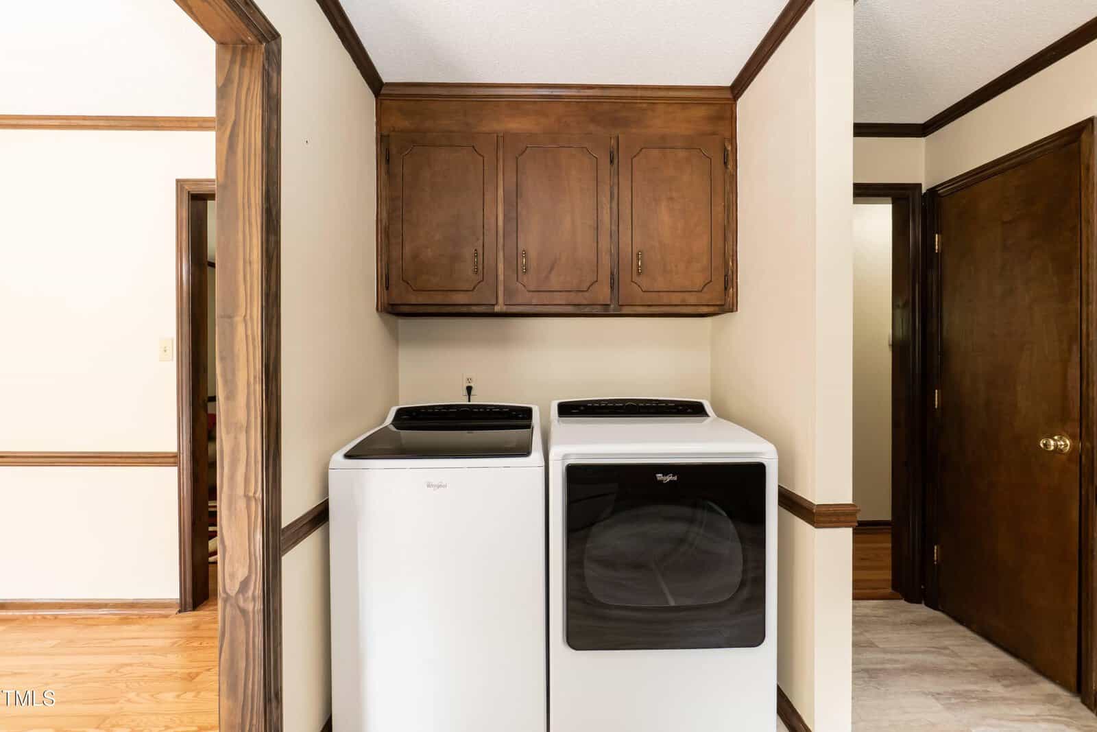 A laundry area with a white washer and dryer below wooden cabinets, set against cream walls with dark wood trim. Doorways and hallways are visible on either side.