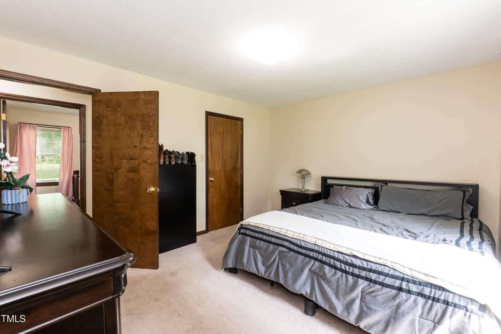A bedroom with a bed, gray bedding, two nightstands, a black dresser, and two wooden doors. There’s a window with pink curtains and a potted plant visible in the adjacent room.