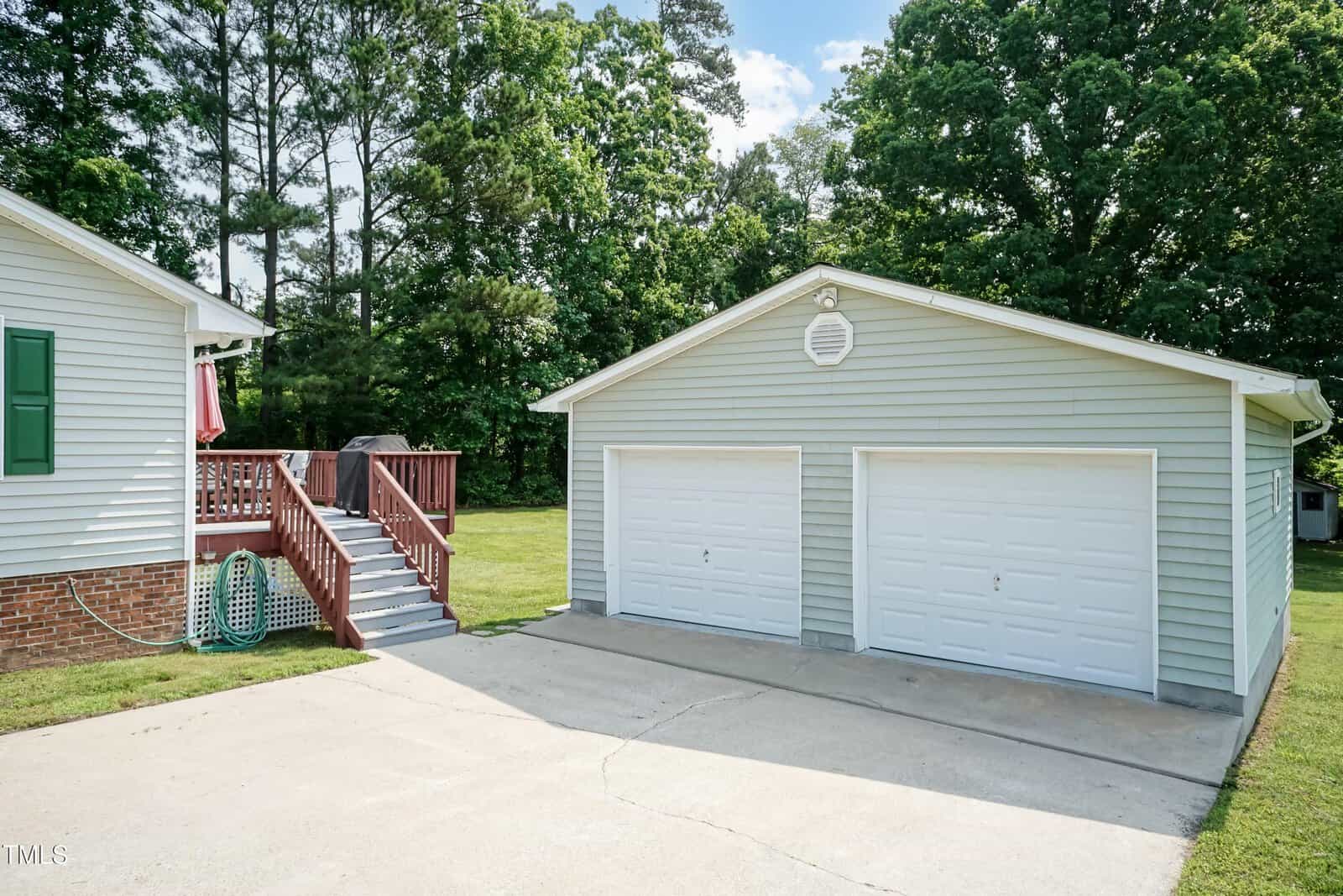 A two-car detached garage with white doors and light gray siding sits next to a house with green shutters and a red staircase, surrounded by trees and a concrete driveway.