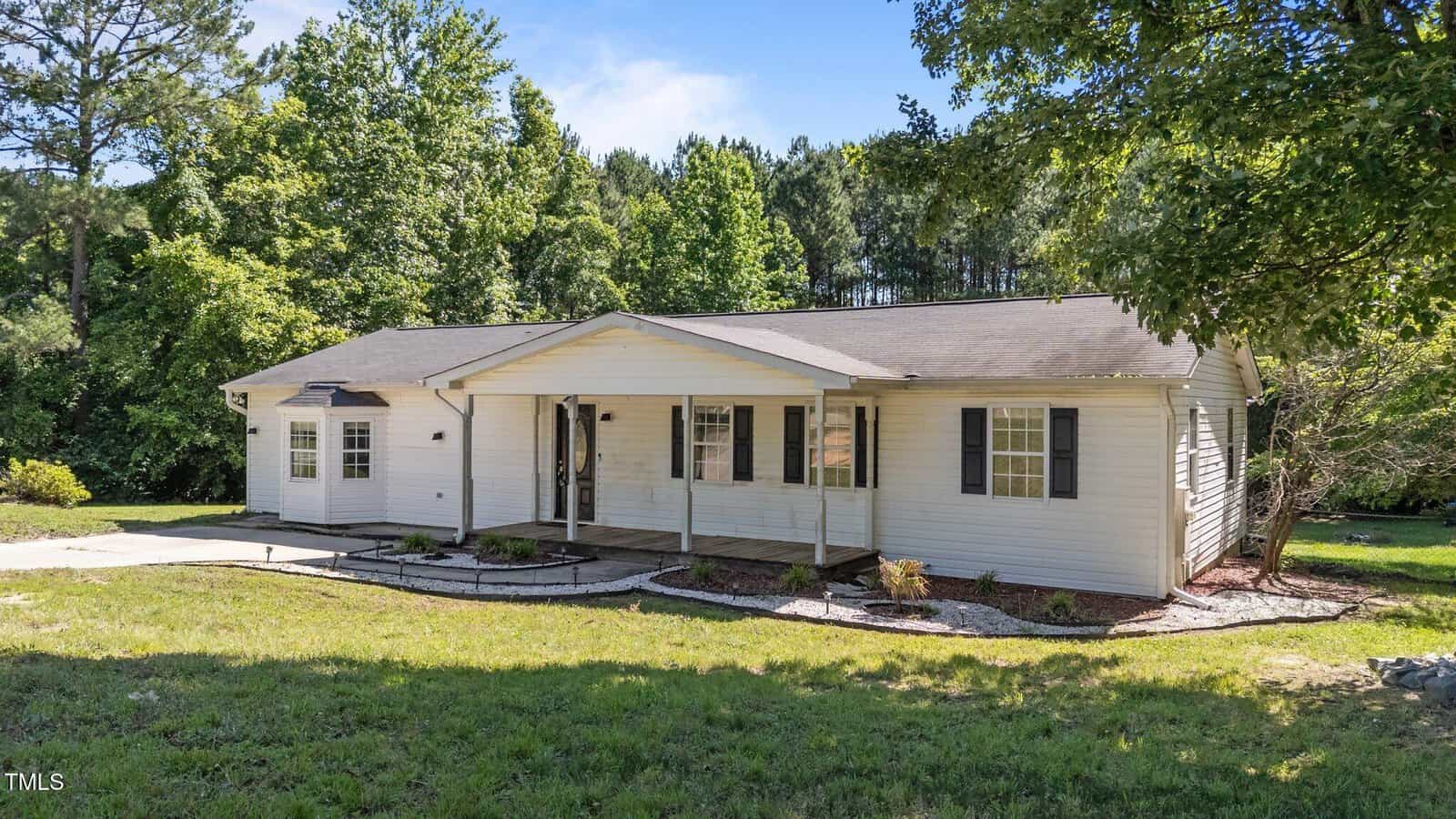 Single-story white house with a covered front porch, black shutters, and a gently sloping roof. Surrounded by green grass, mature trees, and landscaping under a sunny sky.