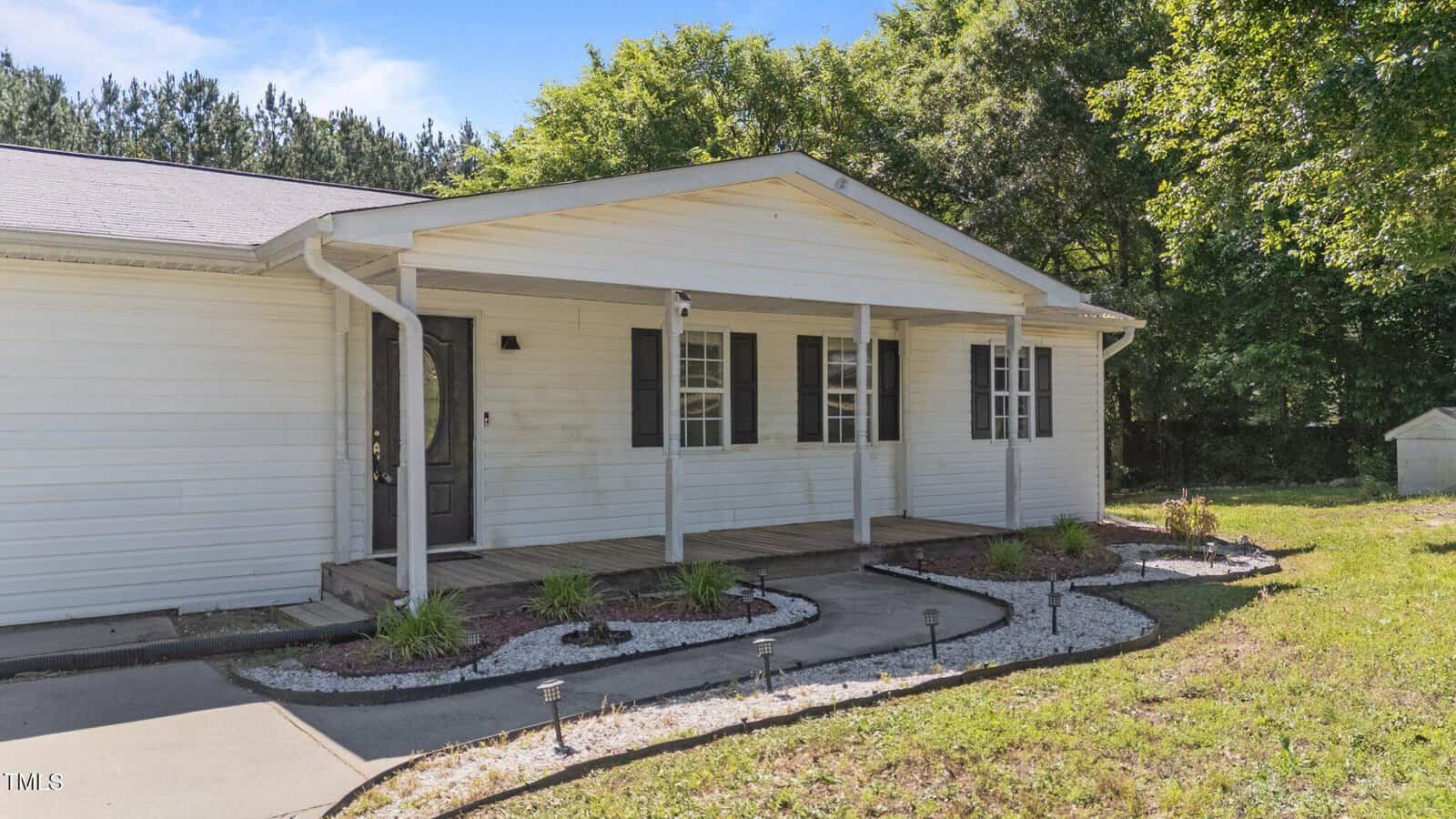 A single-story white house with a covered front porch, black shutters, and a landscaped walkway bordered with white rocks and small plants. Trees and a grassy yard surround the house.