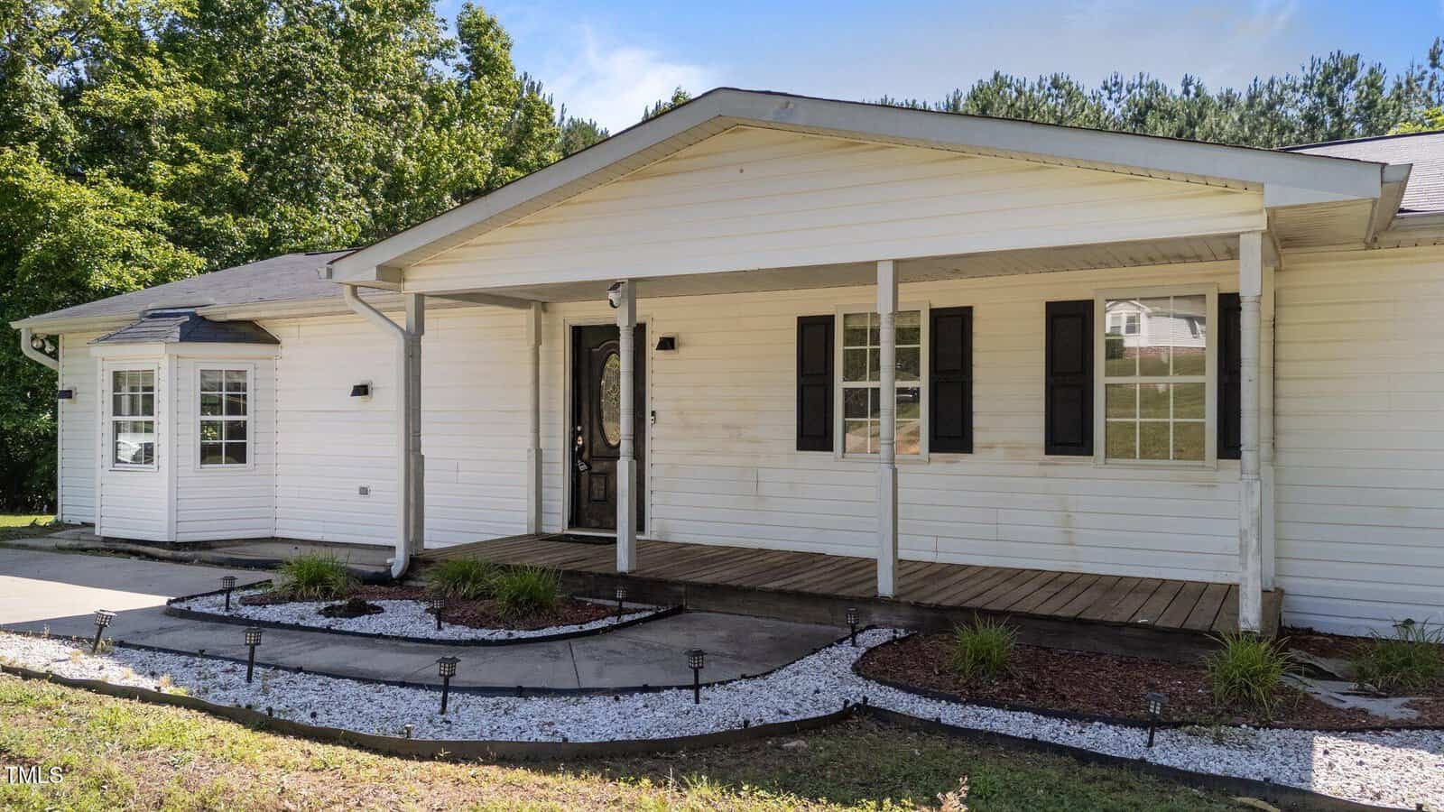 Single-story white house with a covered front porch, black door, and black window shutters. The yard features a curved walkway bordered by white gravel, mulch, and small plants, with trees in the background.
