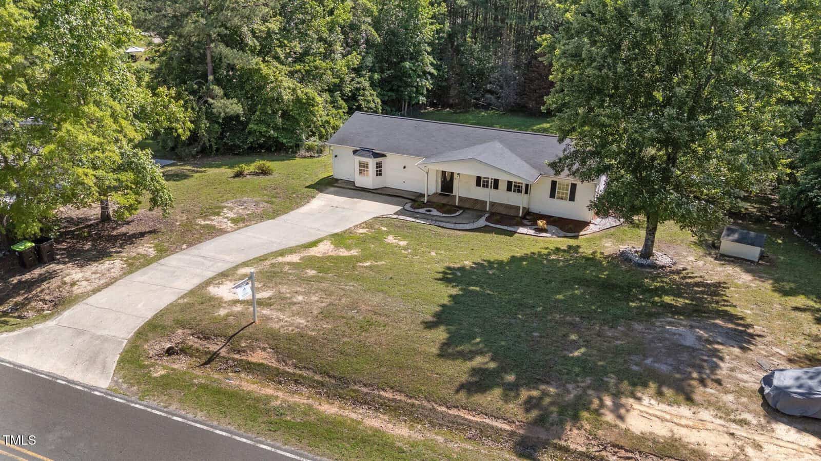 Single-story house with a covered front porch, white exterior, and dark roof, situated on a spacious lot with a curved driveway, large tree in the yard, and surrounded by trees and greenery.